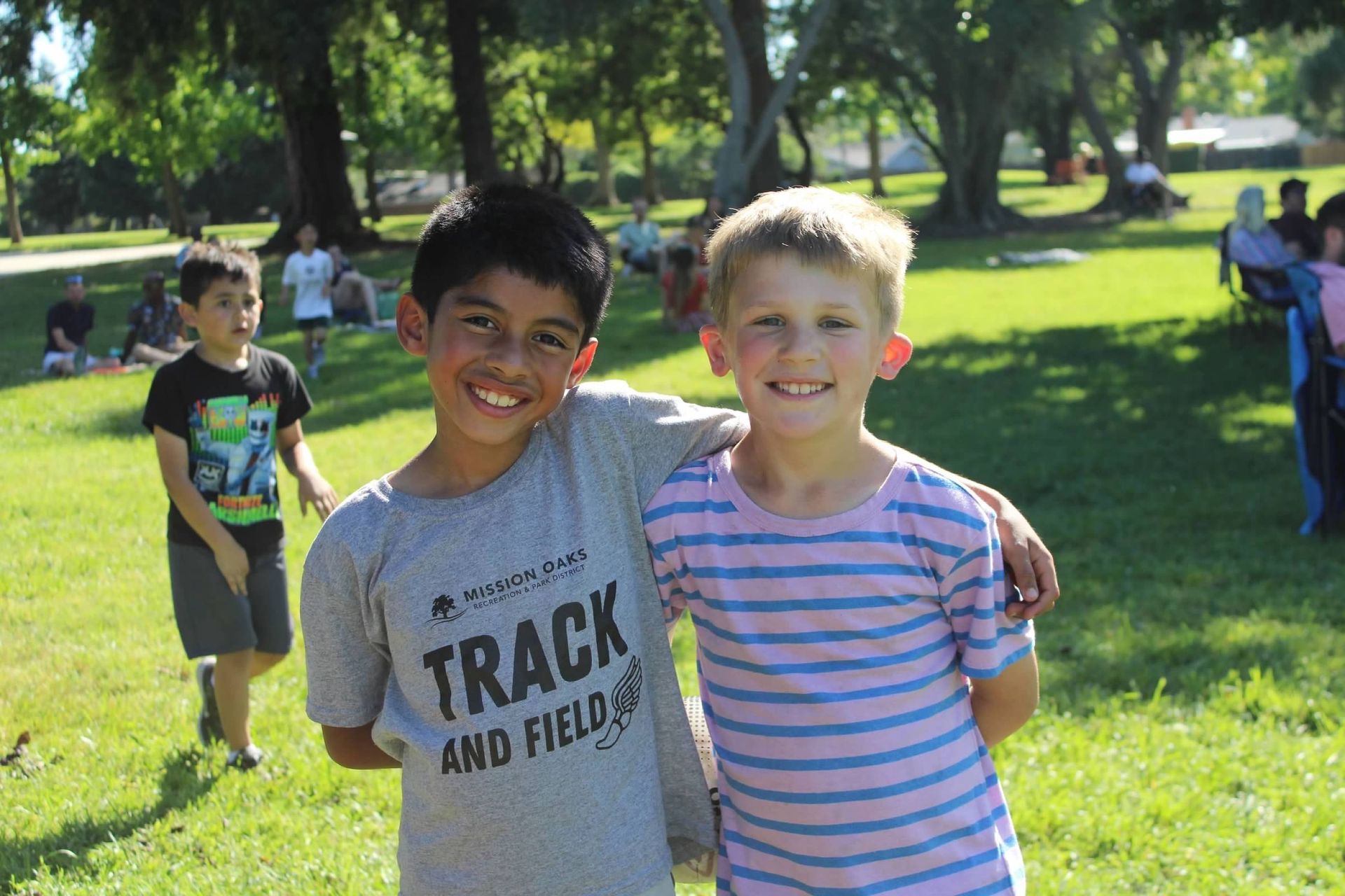 Two boys with arms around each other, smiling outdoors. One in a track shirt, other in blue and pink stripes.
