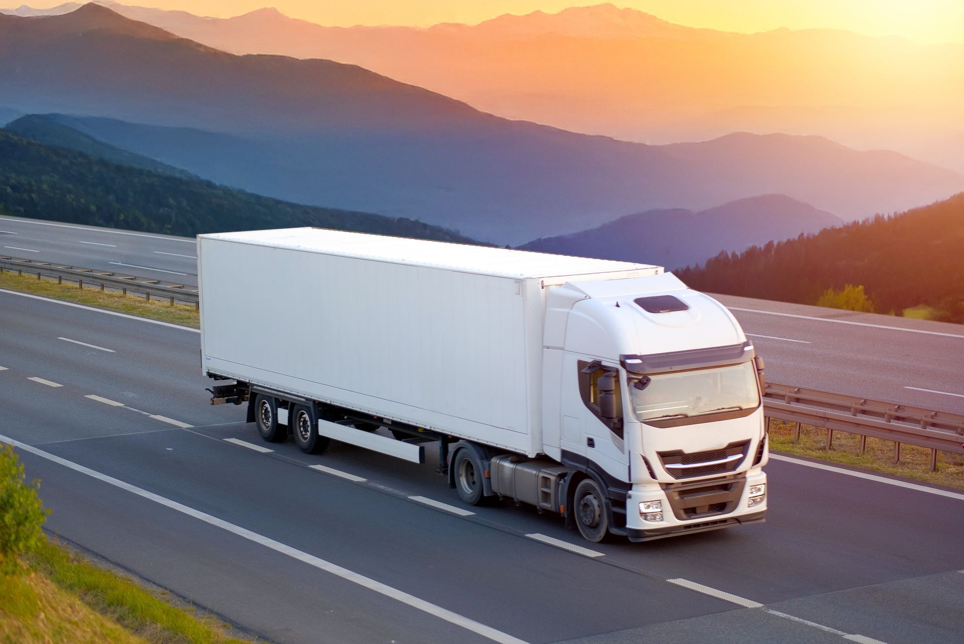 White semi-truck driving on a highway with mountain backdrop at sunset.