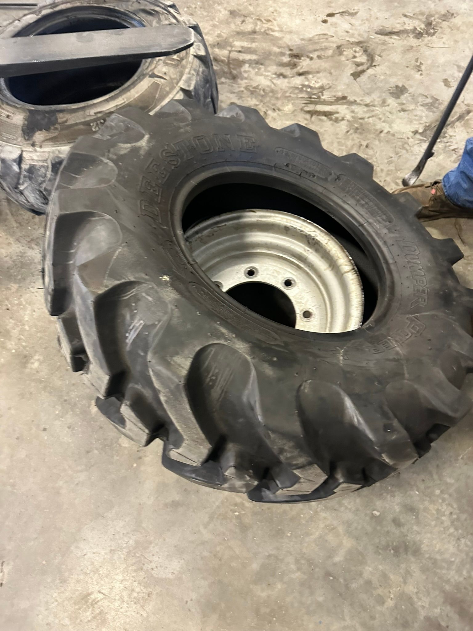 Black tractor tire with silver wheel; another tire is in background.