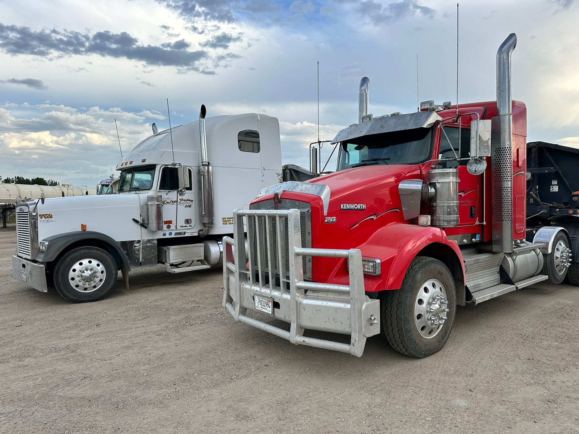 Two semi-trucks, one white, one red, parked outdoors on a gravel lot under a cloudy sky.