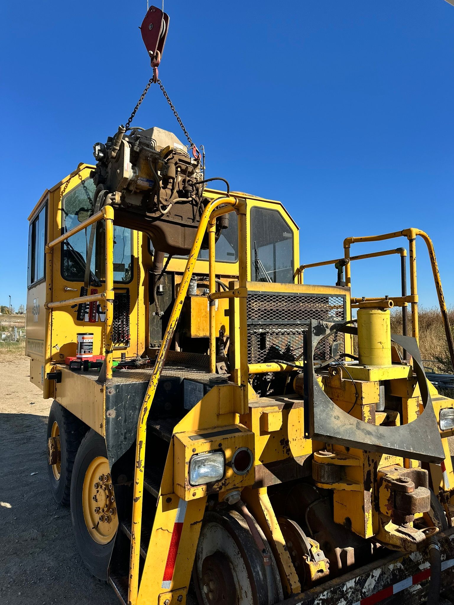 Yellow industrial vehicle with machinery being lifted by a crane against a blue sky.
