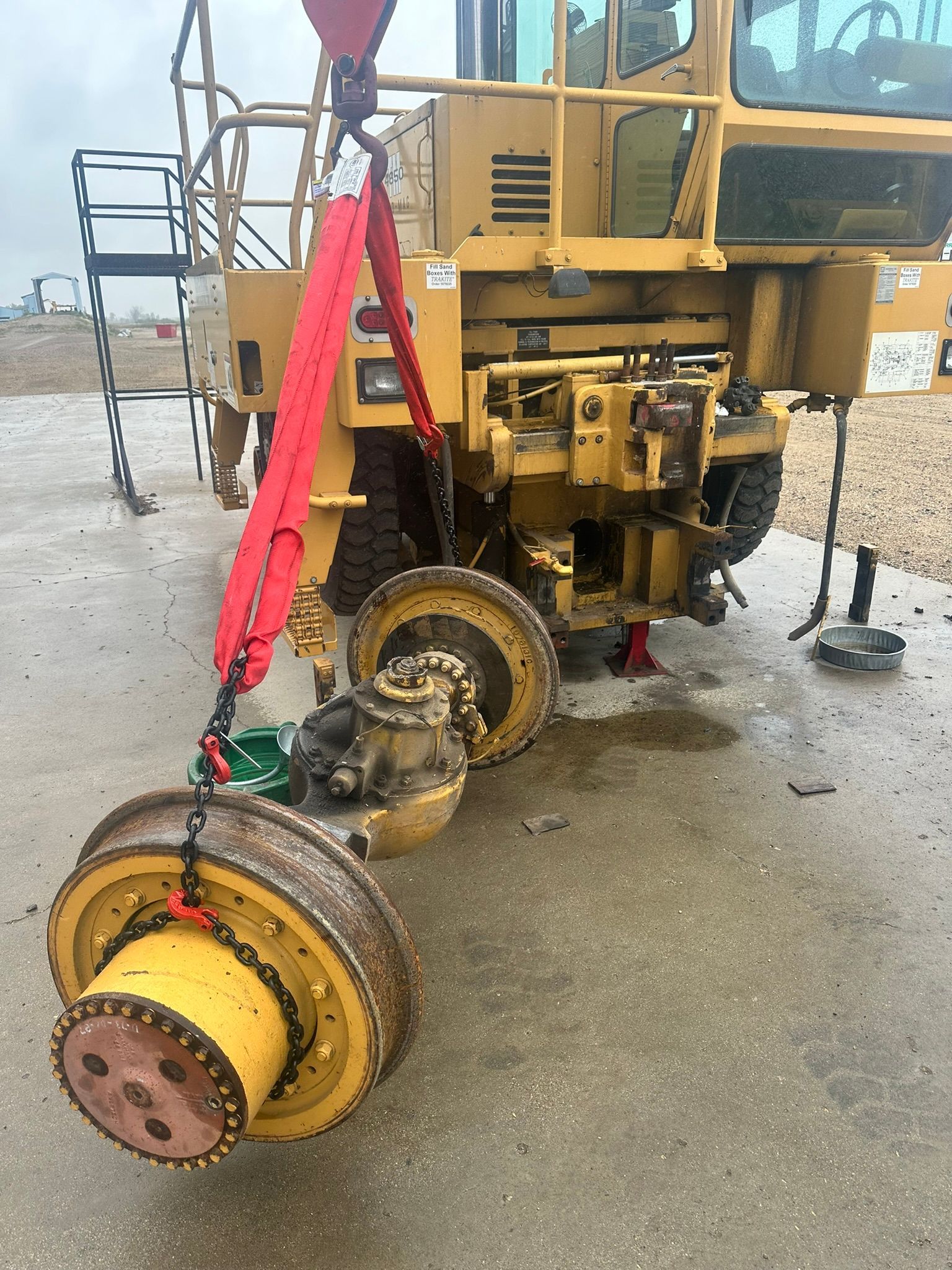 Yellow construction vehicle, rear wheel removed and suspended by a red strap, outdoors.