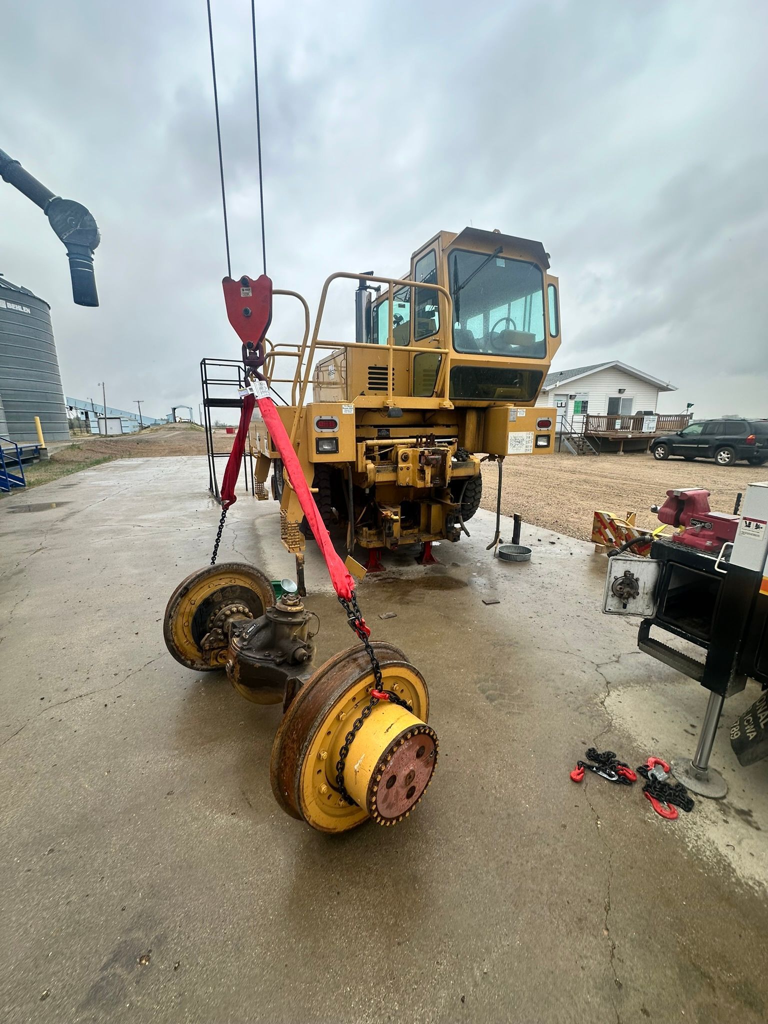A yellow industrial machine with removed wheels being lifted by a crane outdoors on a wet surface.