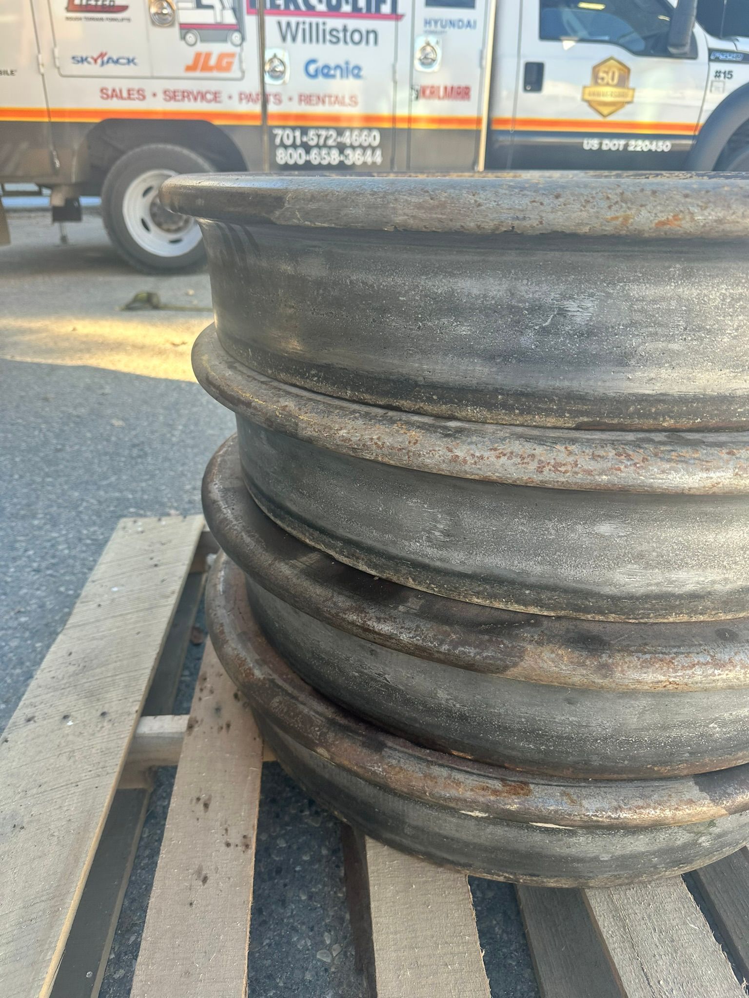 Stack of dark, rusty metal rims on a wooden pallet, with a white truck in the background.