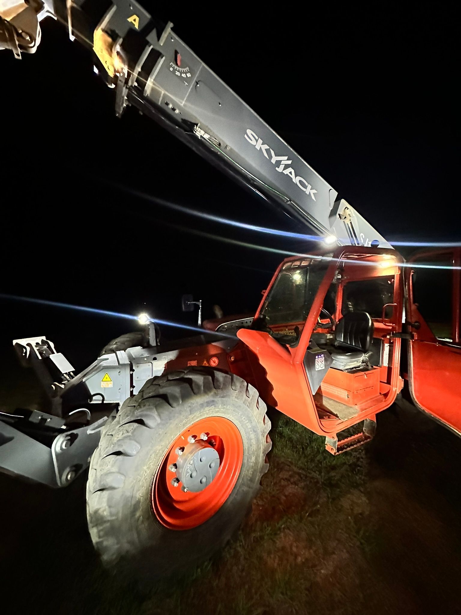 Orange Skyjack telehandler at night, lights on, boom extended.
