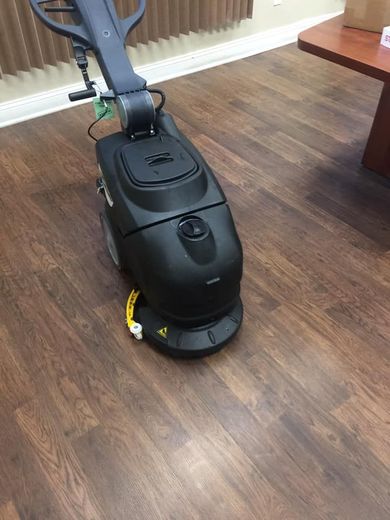 A black automatic floor scrubber sitting on a wood-look floor near a brown table.
