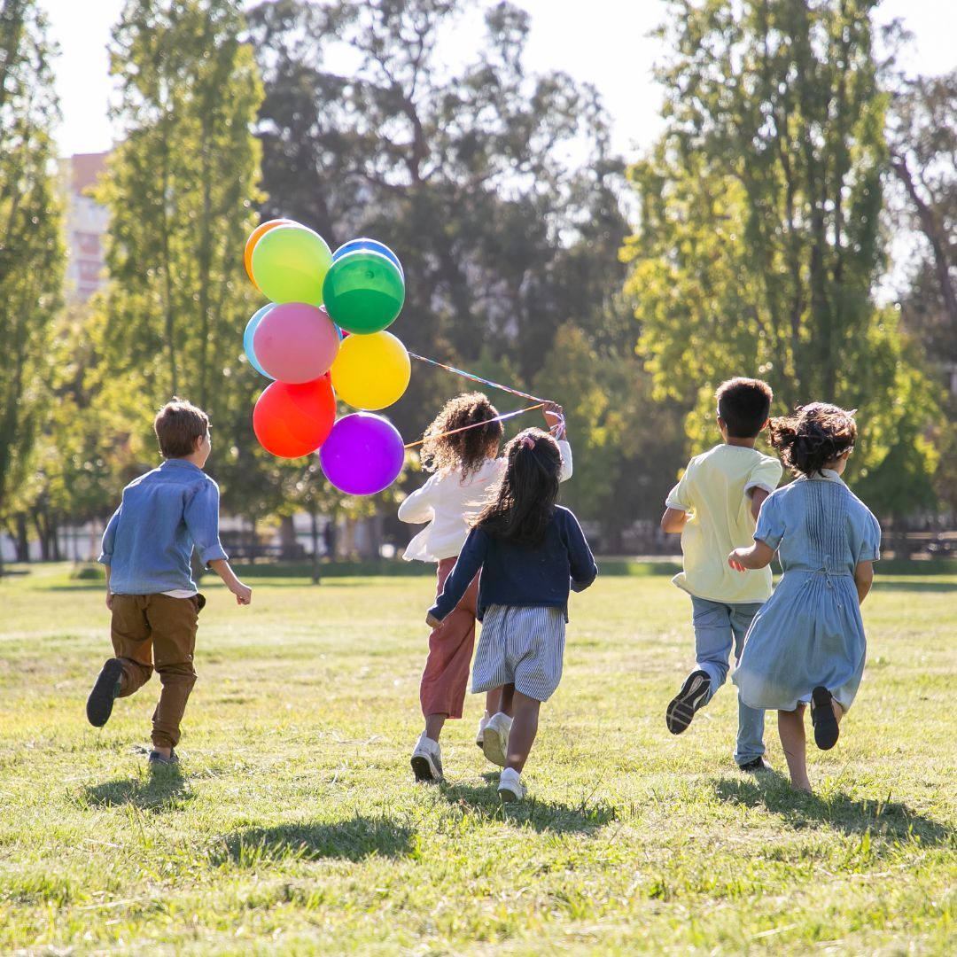 A Courageous Voice / A group of five children running in a grassy field on a sunny day. One girl in the group is holding a bundle of various colored balloons. 