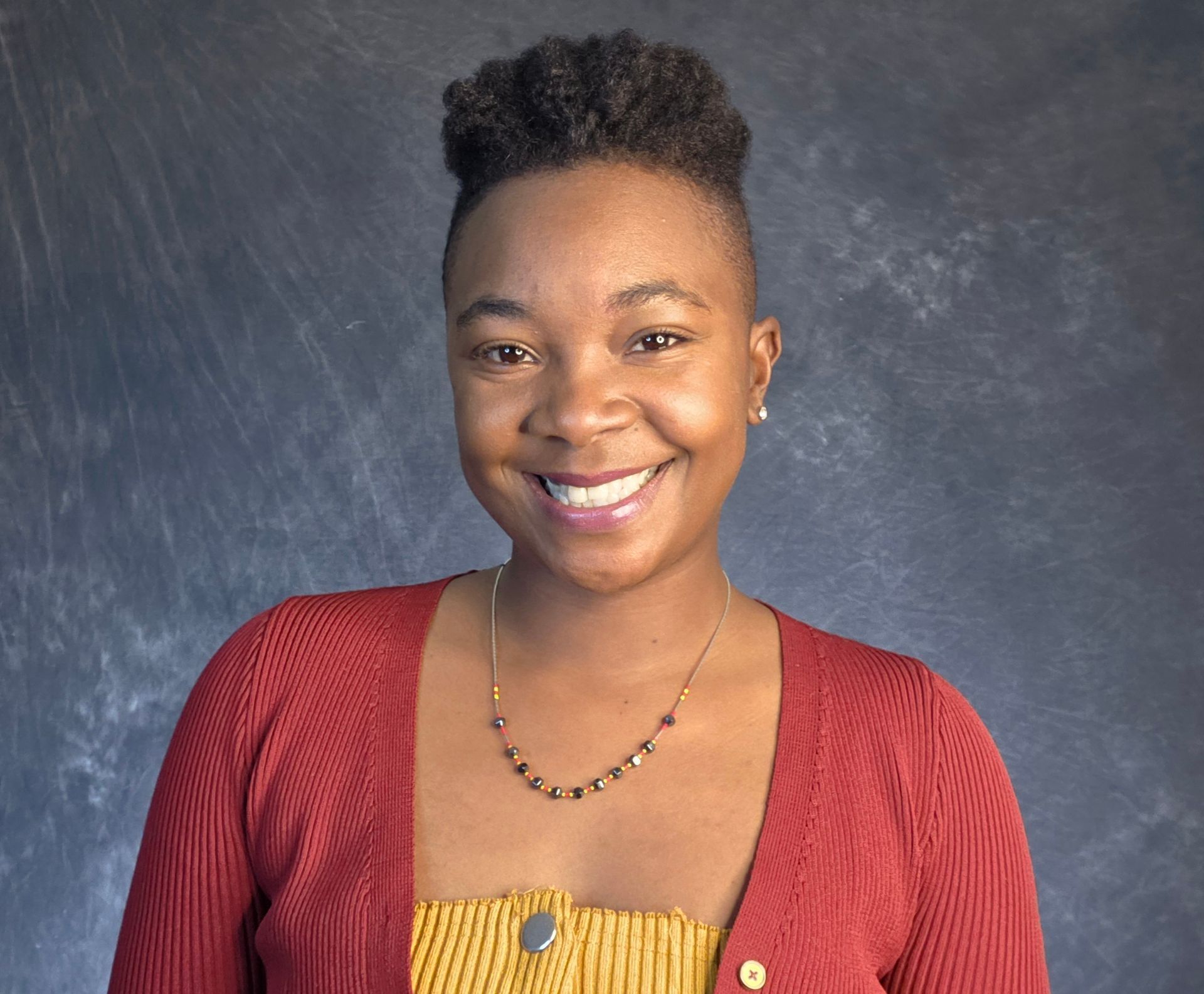 Smiling person in red sweater and yellow top, dark hair, against a textured gray background.