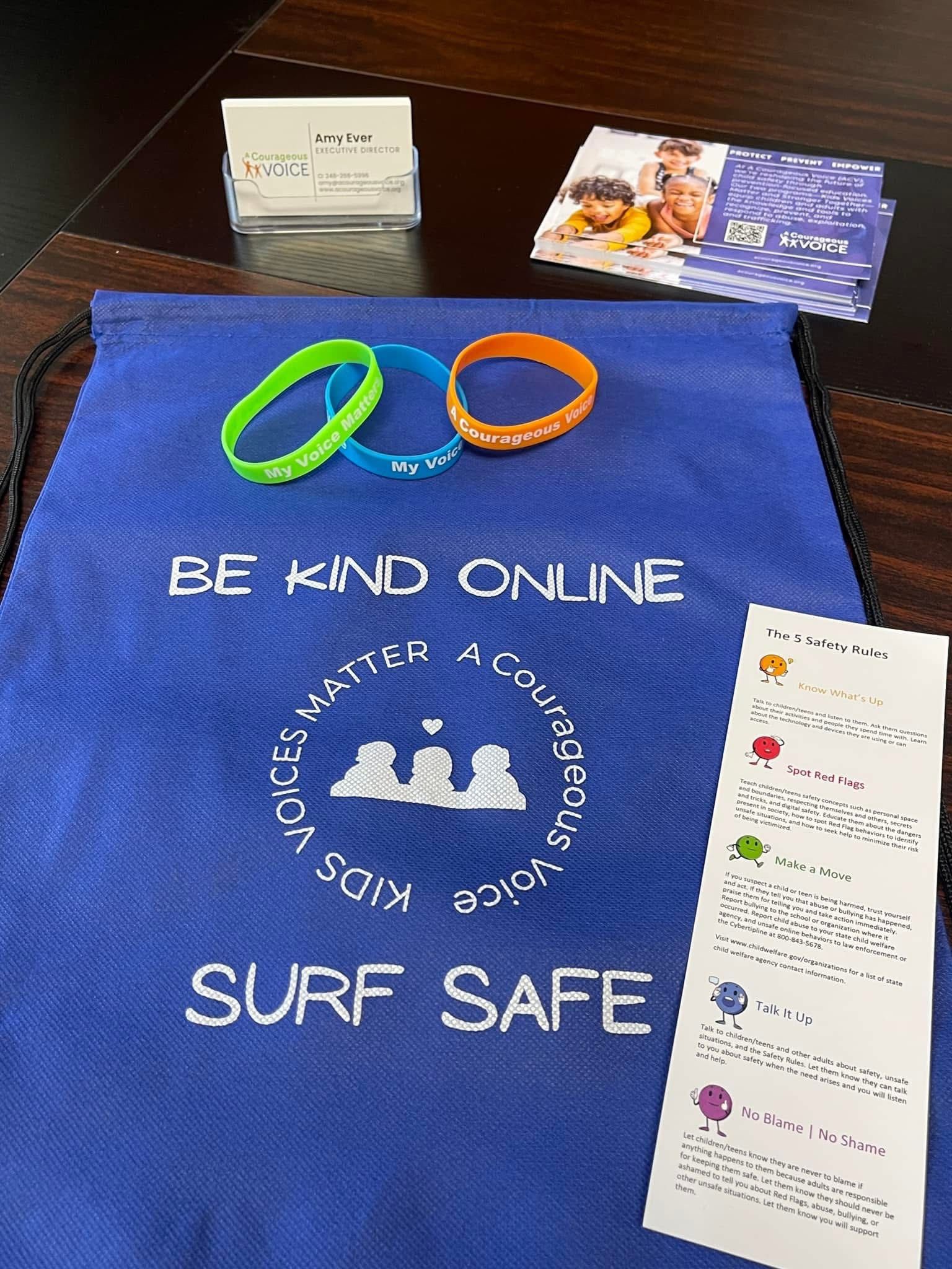 Blue tote with “BE KIND ONLINE” and “SURF SAFE,” plus two wristbands and info cards on a table.