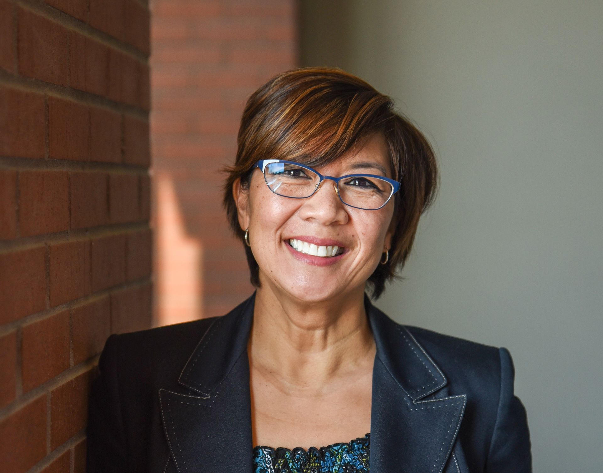 Woman smiling, wearing glasses and a blazer, leaning against a brick wall.