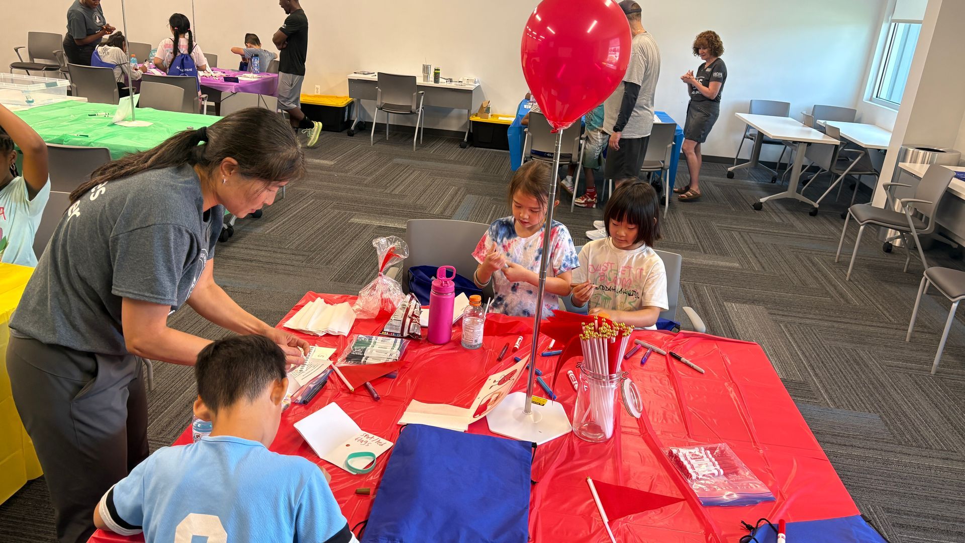 People gathered around a red-covered activity table with balloons at an indoor event.