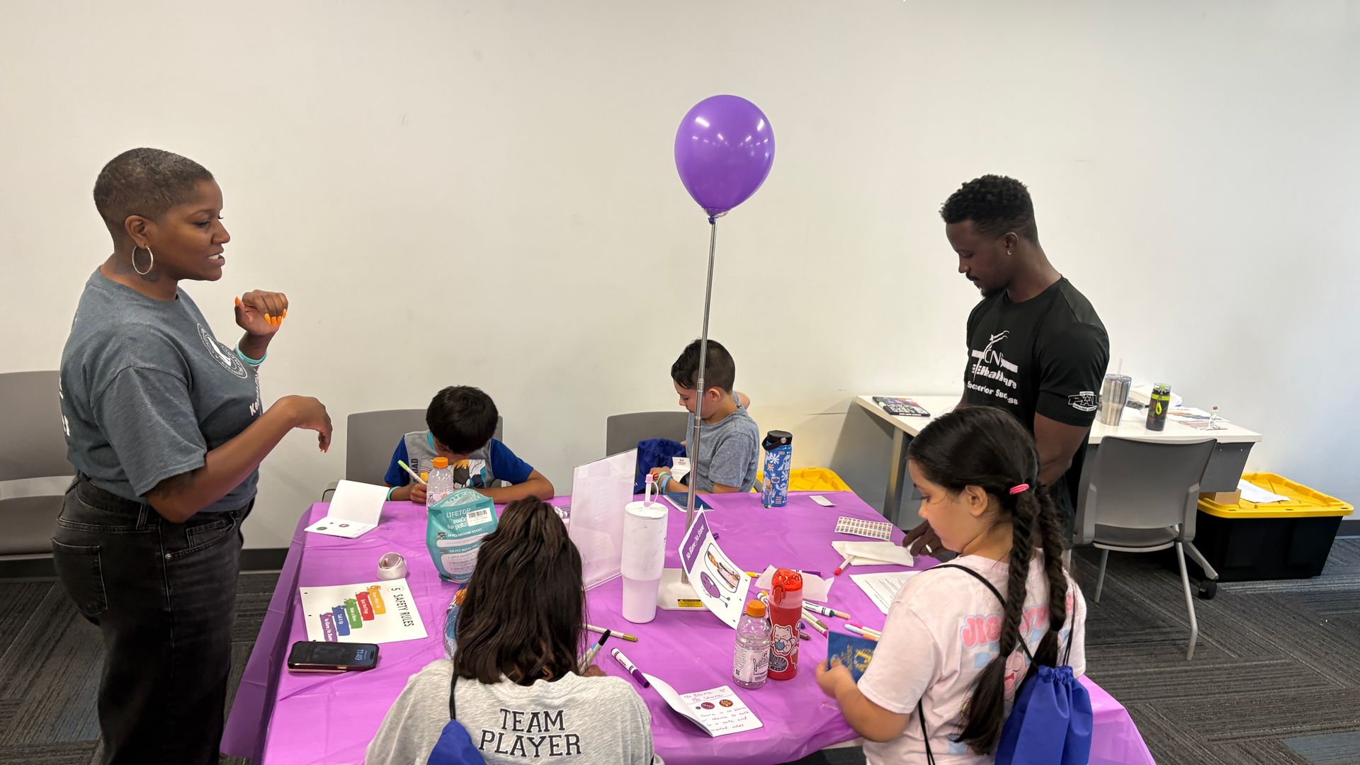 People seated around a purple-covered table at a small birthday party, with a balloon and snacks in a room.