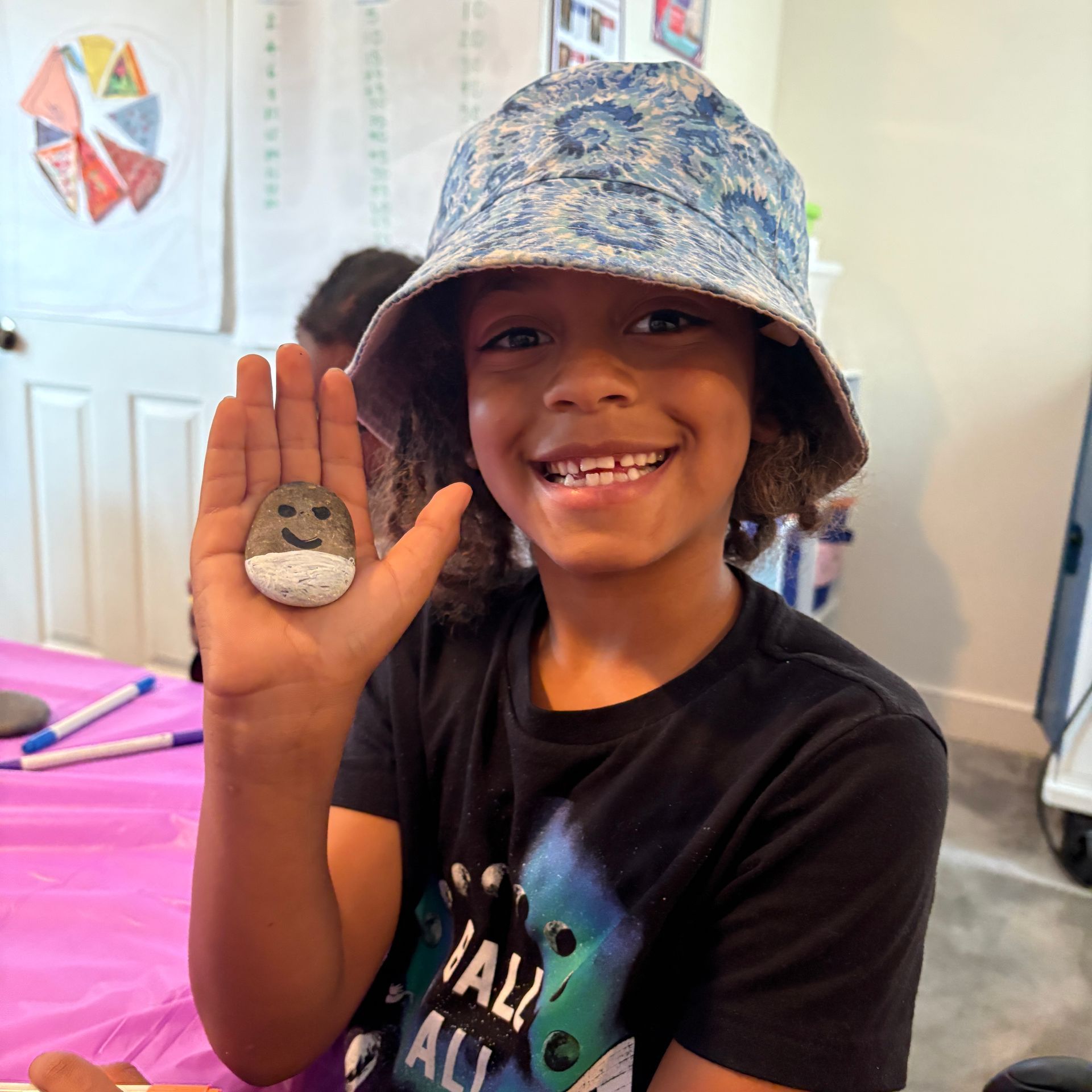 Child smiling and holding up a painted rock while wearing a blue patterned bucket hat indoors