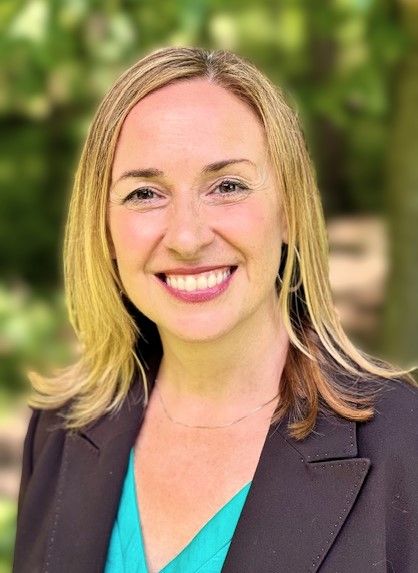 Woman with blonde hair, smiling in a brown blazer and teal top, standing outside.