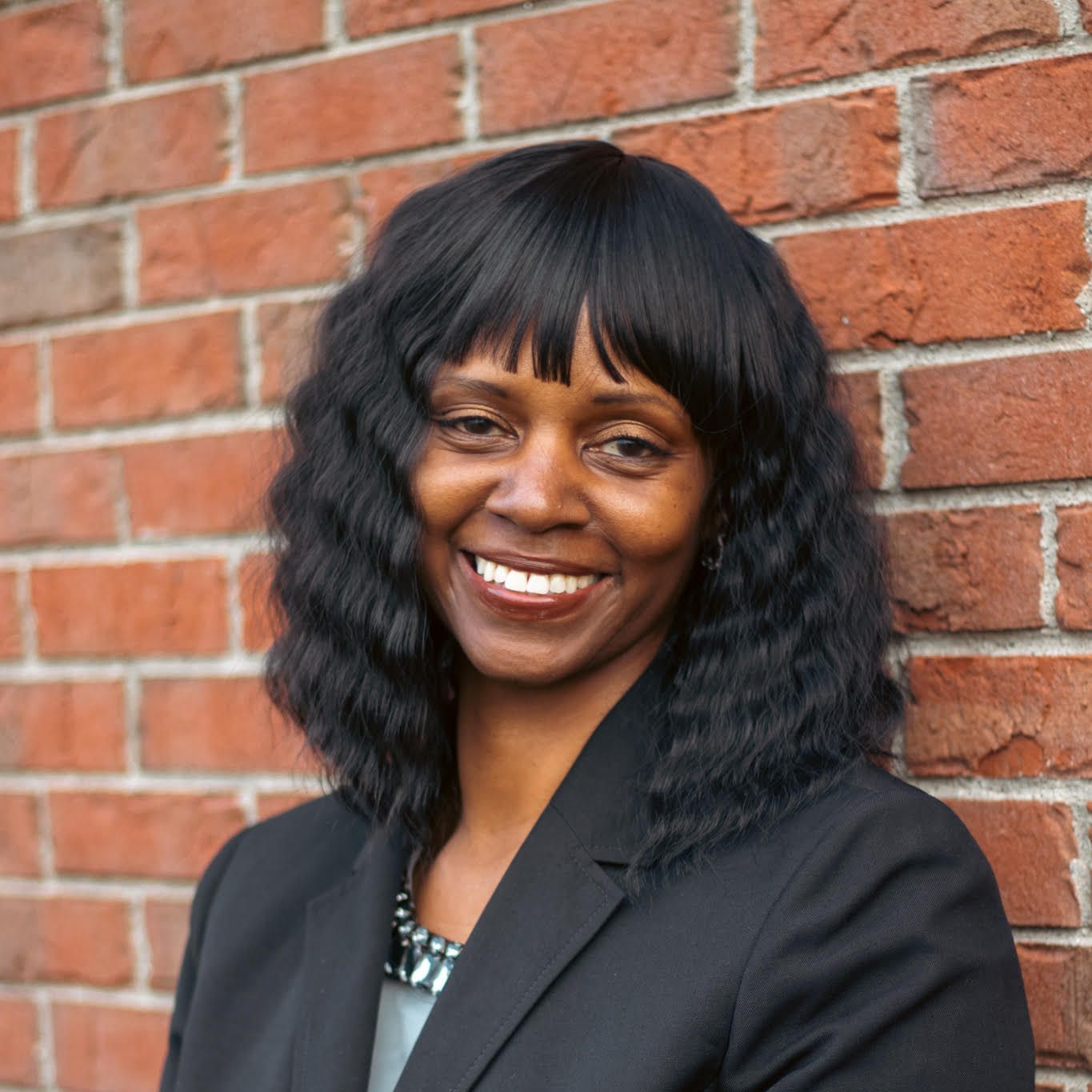 A woman in a black suit is smiling in front of a brick wall