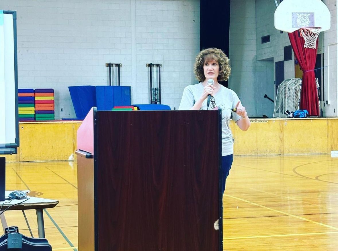 A woman is standing at a podium in a gym talking into a microphone.