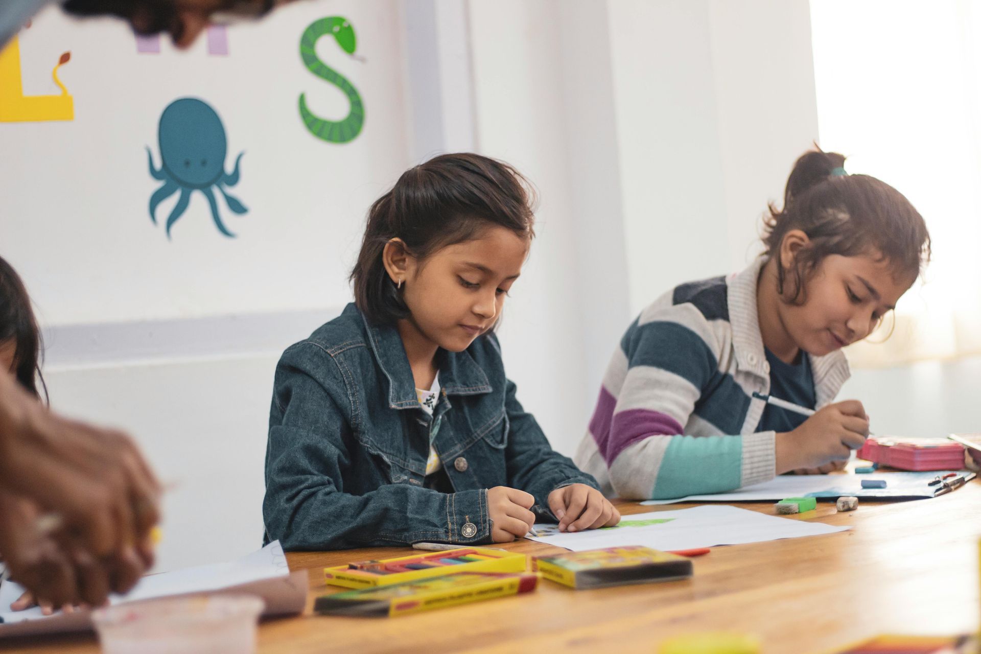 Two children sitting at a table in a classroom drawing with crayons, with an instructor's hands visible in the foreground.