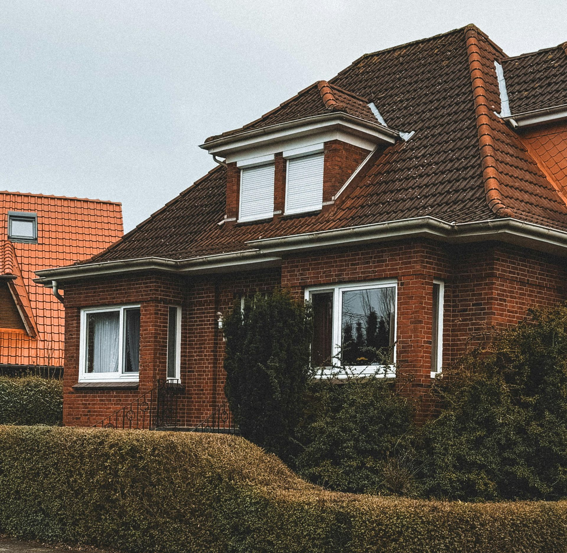 Red brick house with a red tiled roof and white windows, partly hidden by hedges.