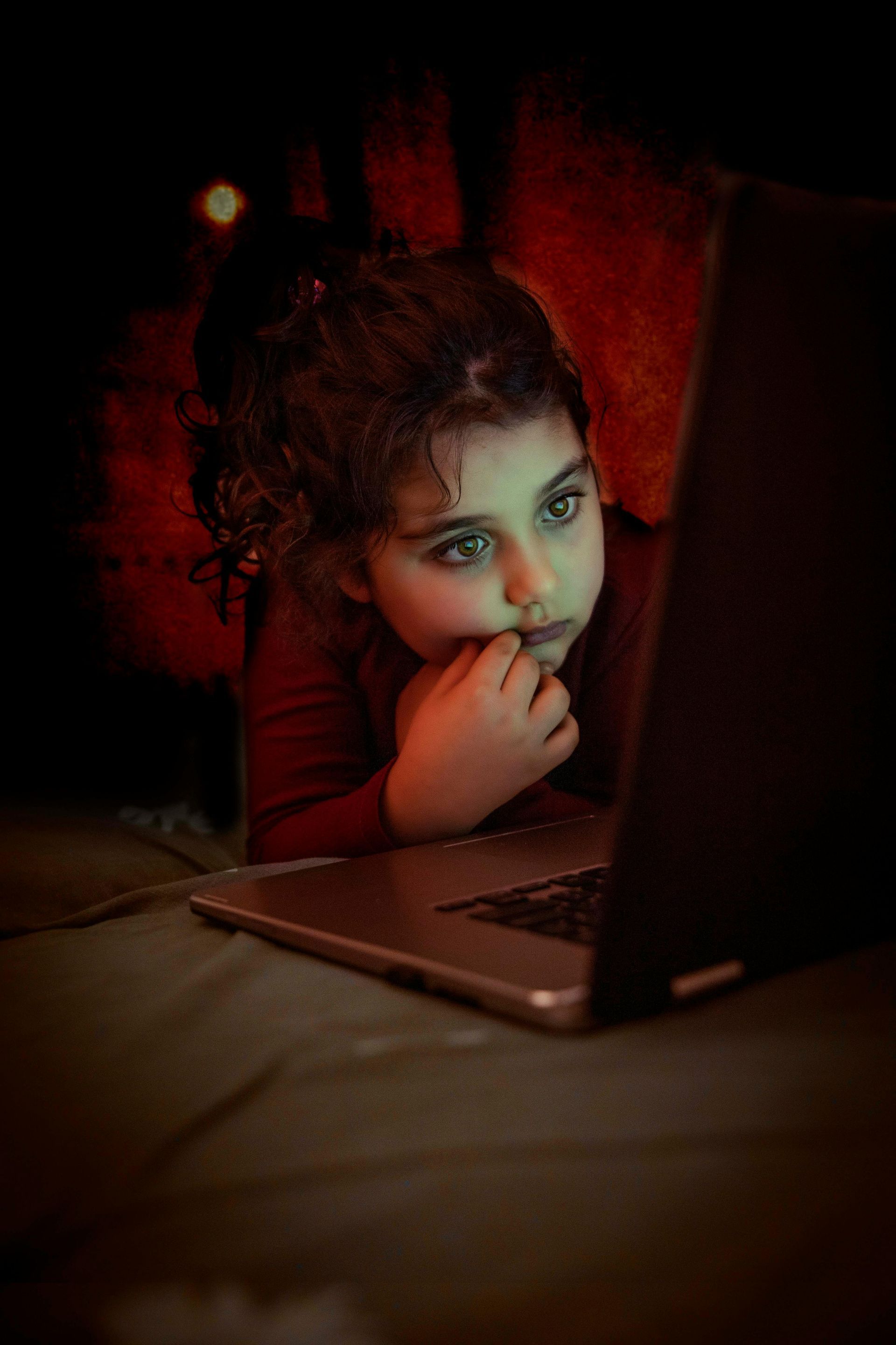 Girl with curly hair looks intently at a laptop screen, resting on a bed. Dark room, red shirt.