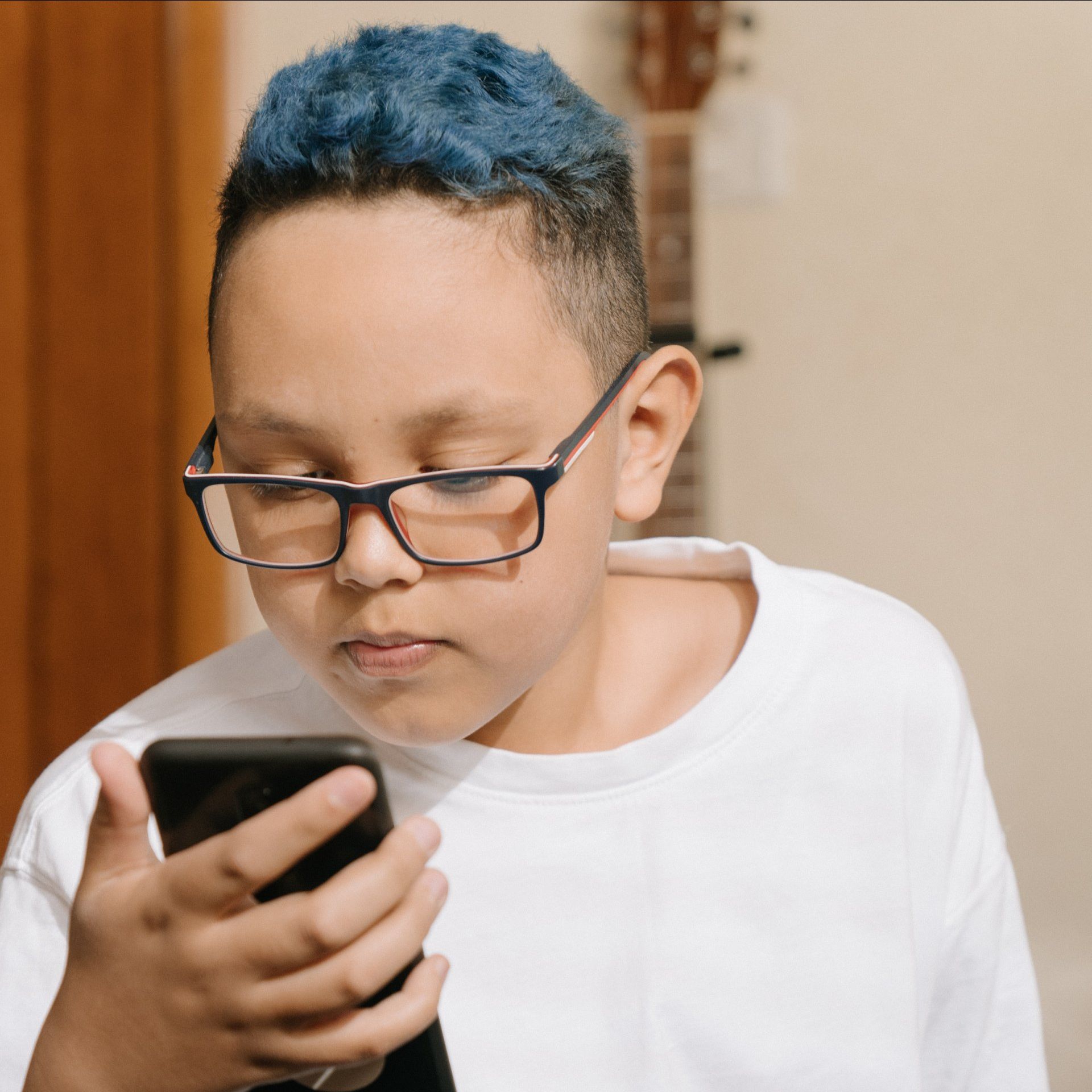 A young boy with blue hair and glasses is looking at a cell phone