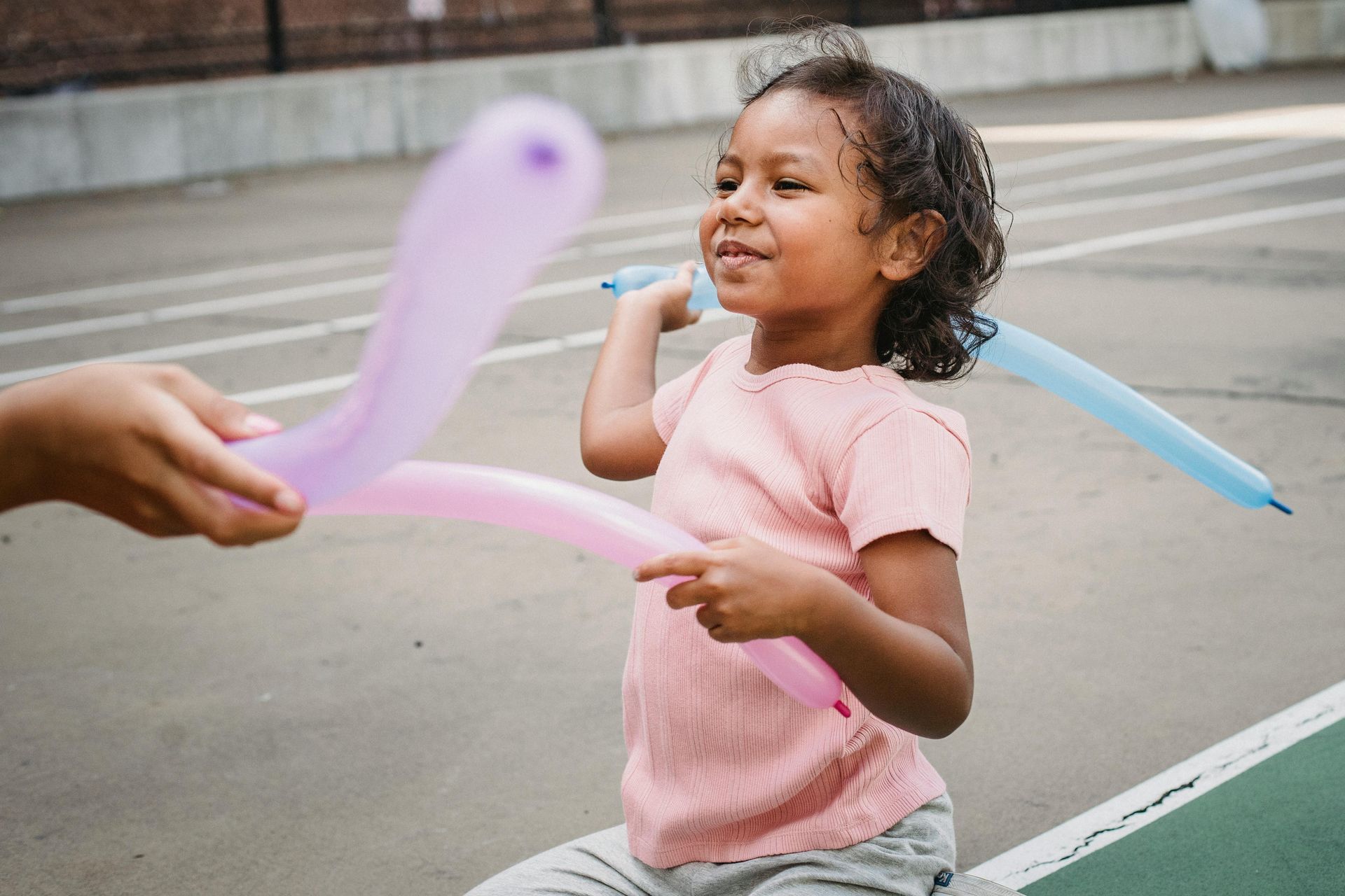 Child in a pink shirt smiling while reaching for a balloon animal on a street sidewalk