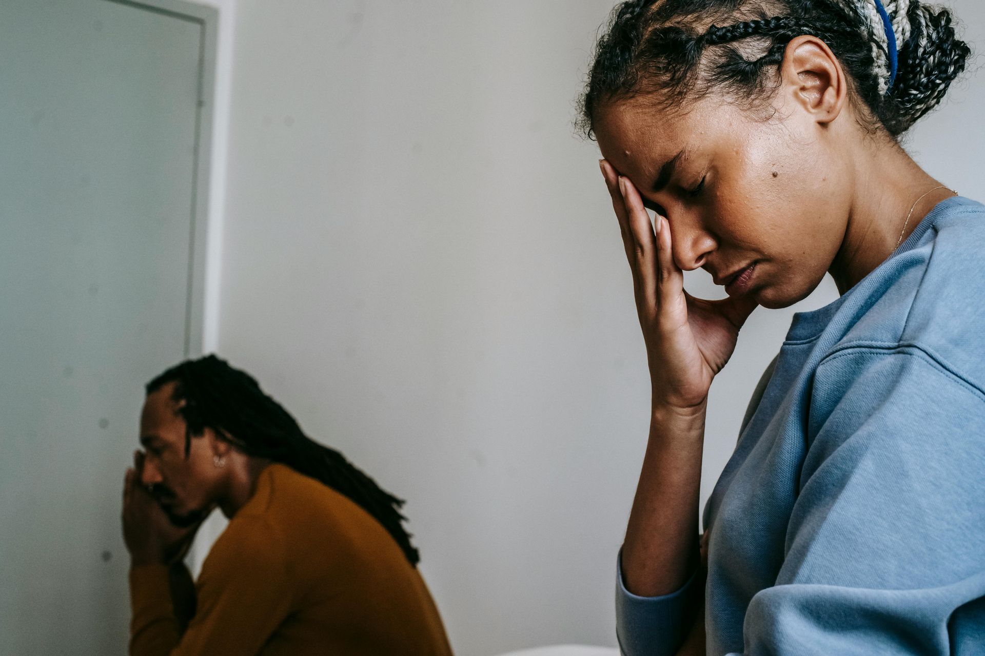 Two people sit apart indoors, each holding a hand to their face in a worried pose.
