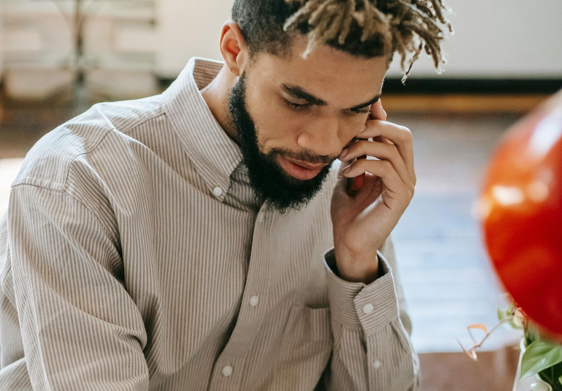 Man with dreadlocks and beard talking on the phone; wearing a striped button-down shirt.