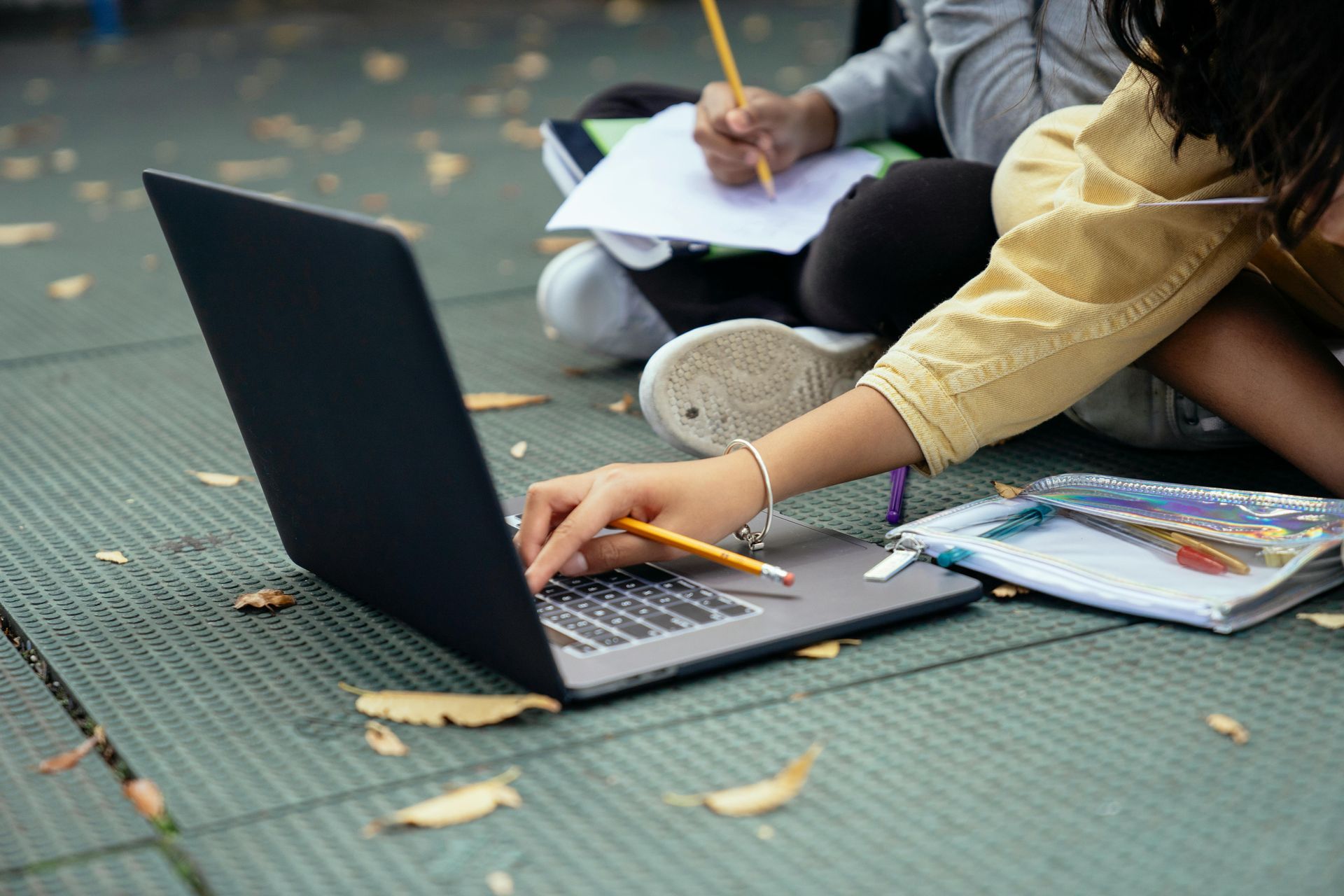 Person using a laptop on the ground, another person writing in a notebook, outdoor setting.