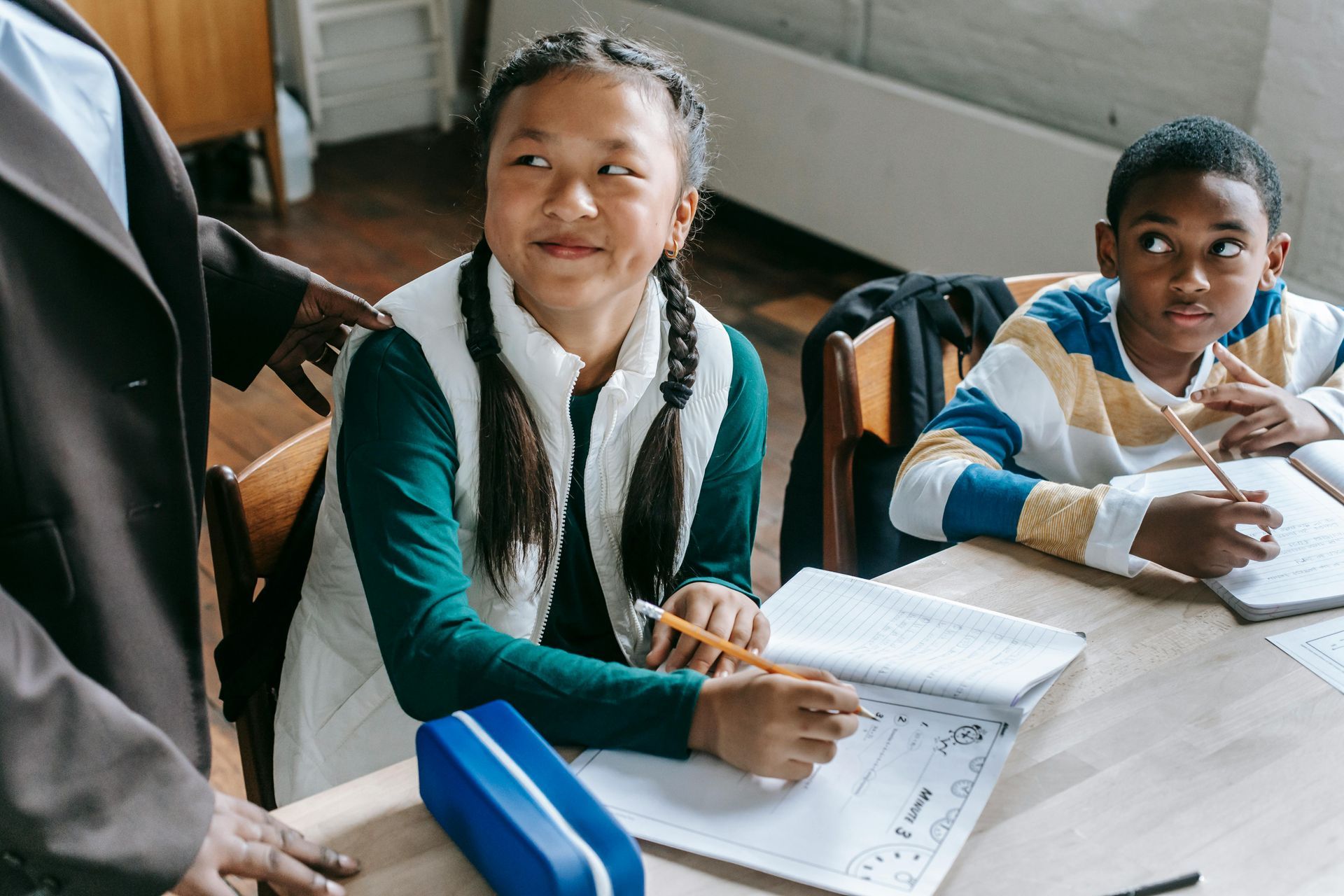 Students at a desk, looking at a teacher. One has a pencil and is writing in a notebook.