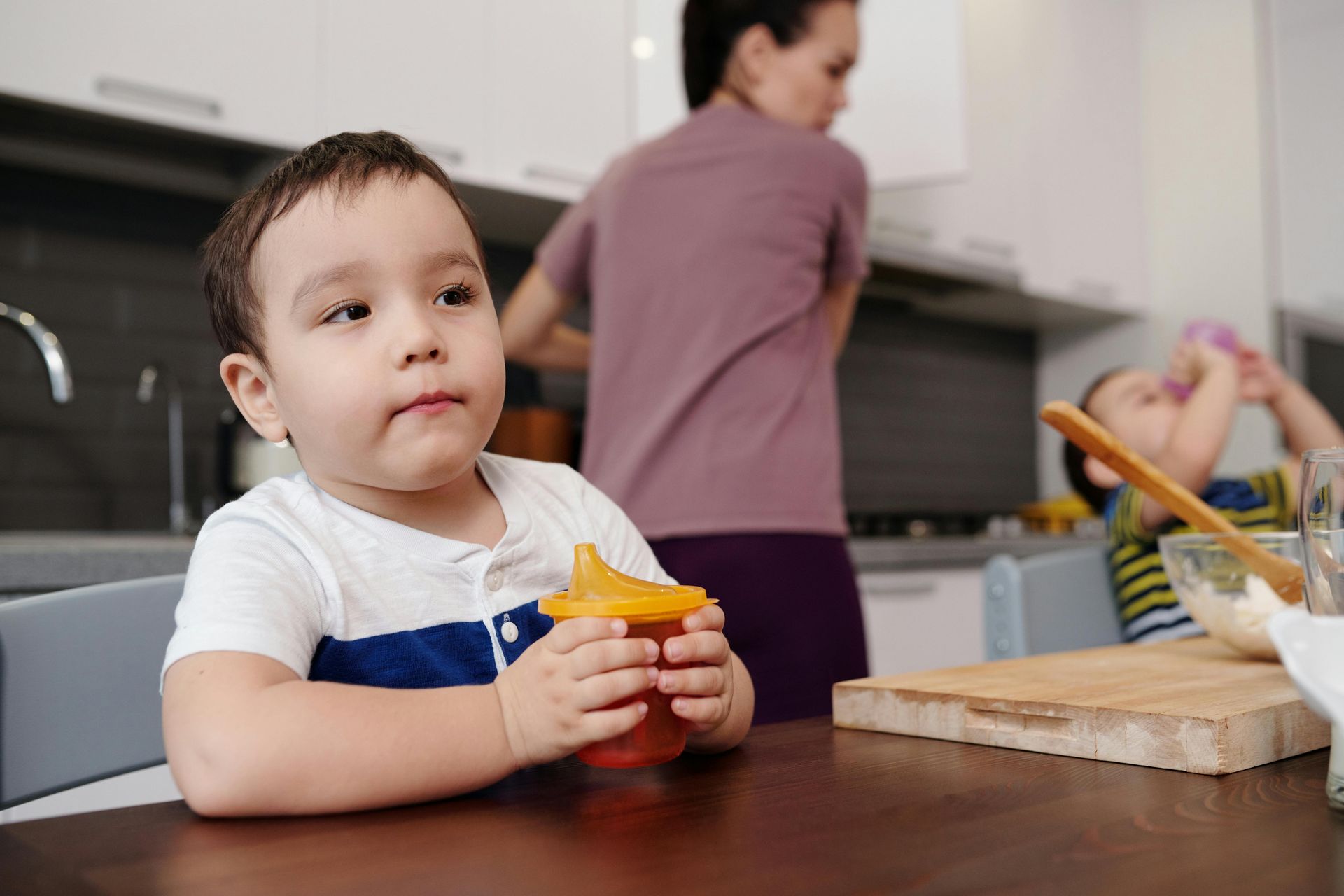 Child sitting at a kitchen table holding a cup, with an adult and another child in the background