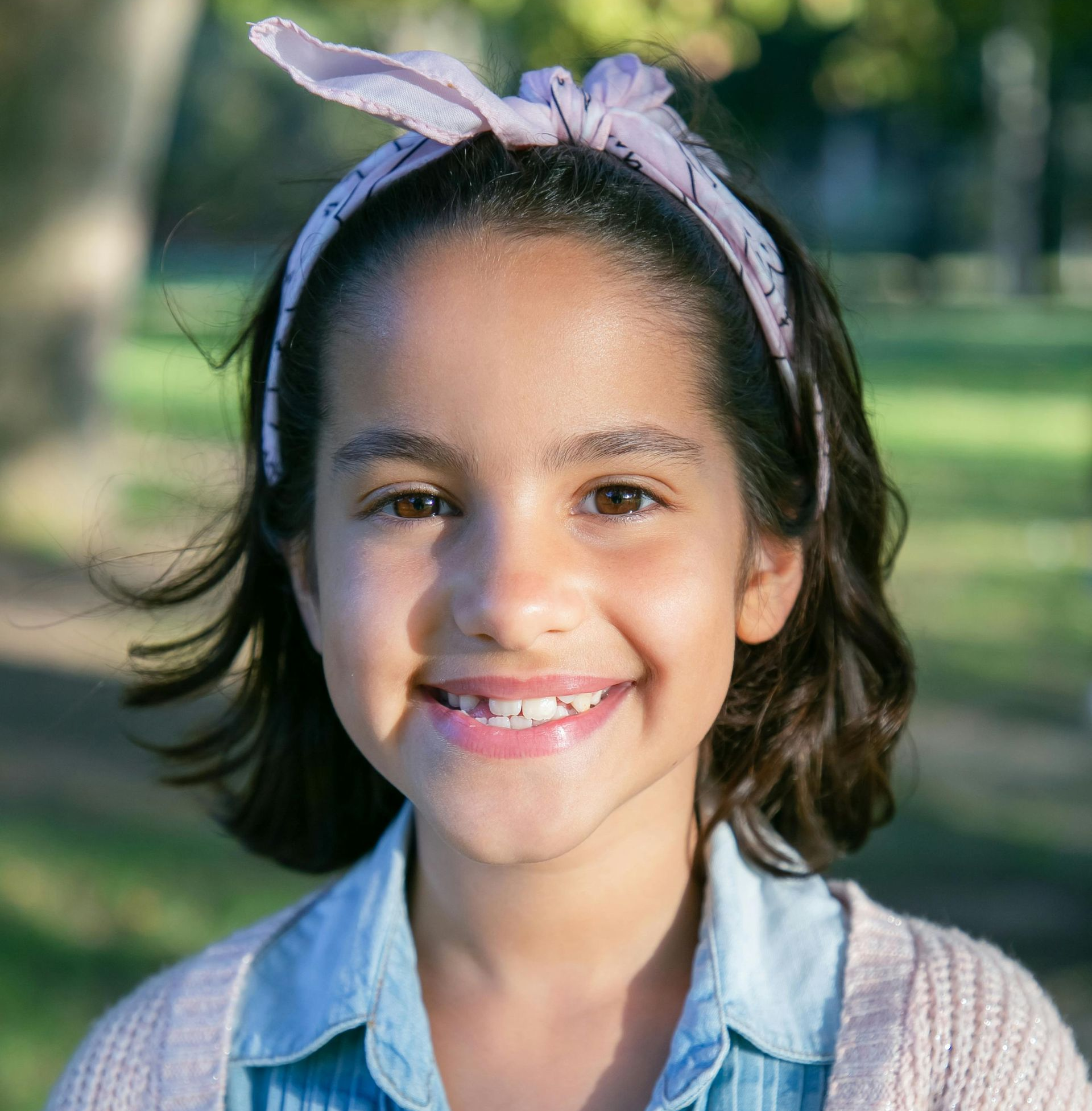 Girl smiling, wearing a blue shirt, pink cardigan, and a headband outdoors.