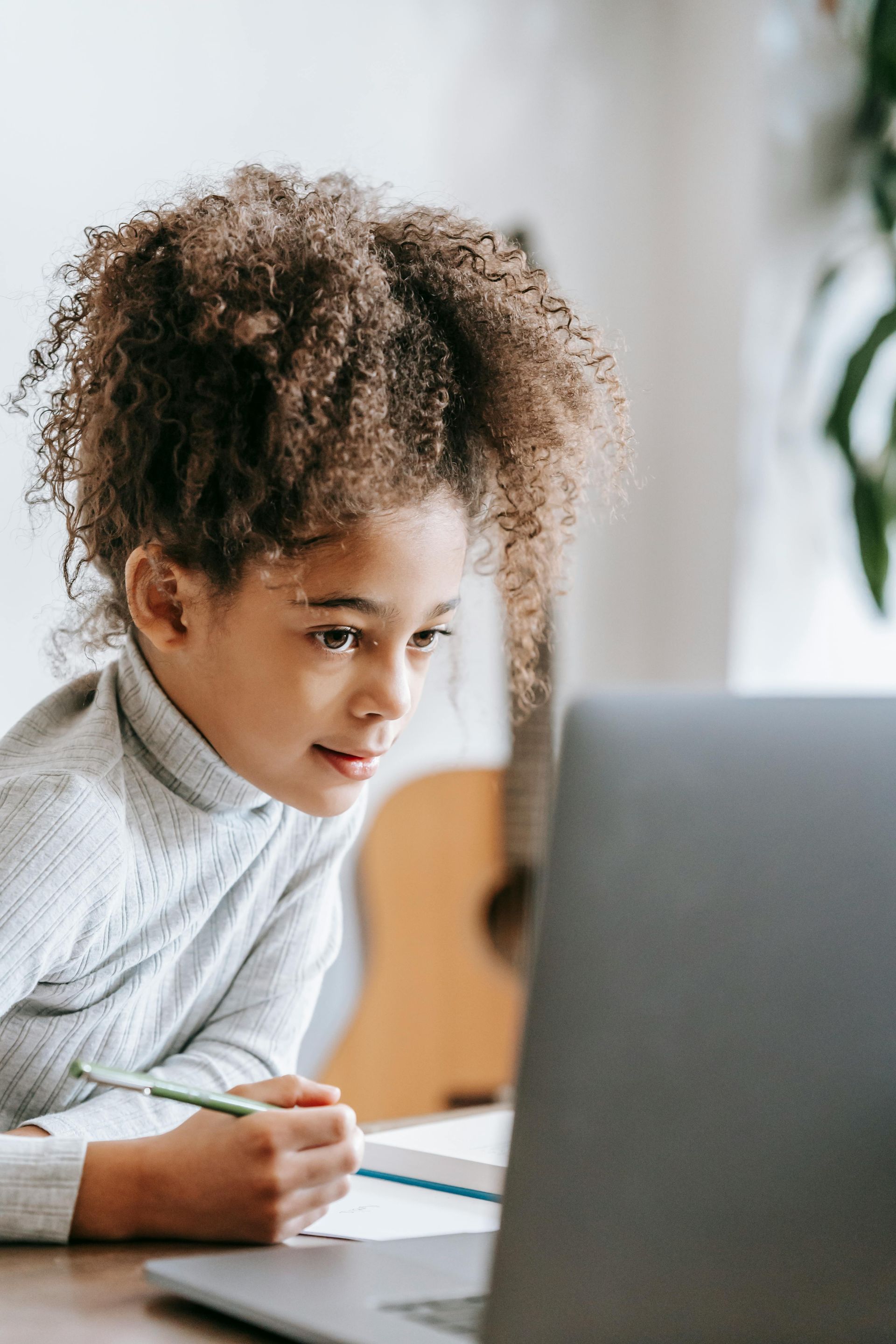 A child with curly hair looks at a laptop while writing with a pencil.