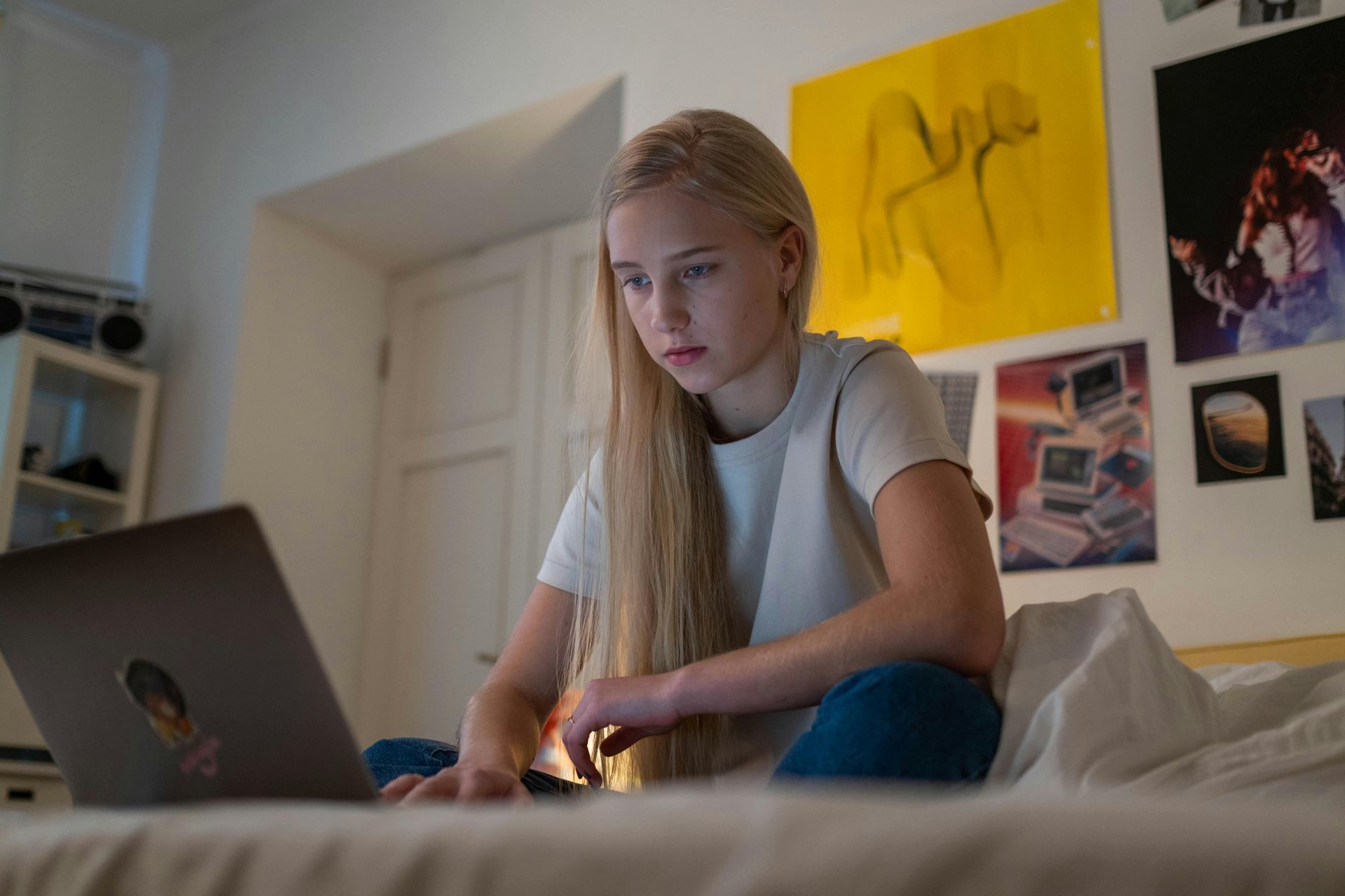 Blond girl using a laptop while sitting on a bed in a bedroom.