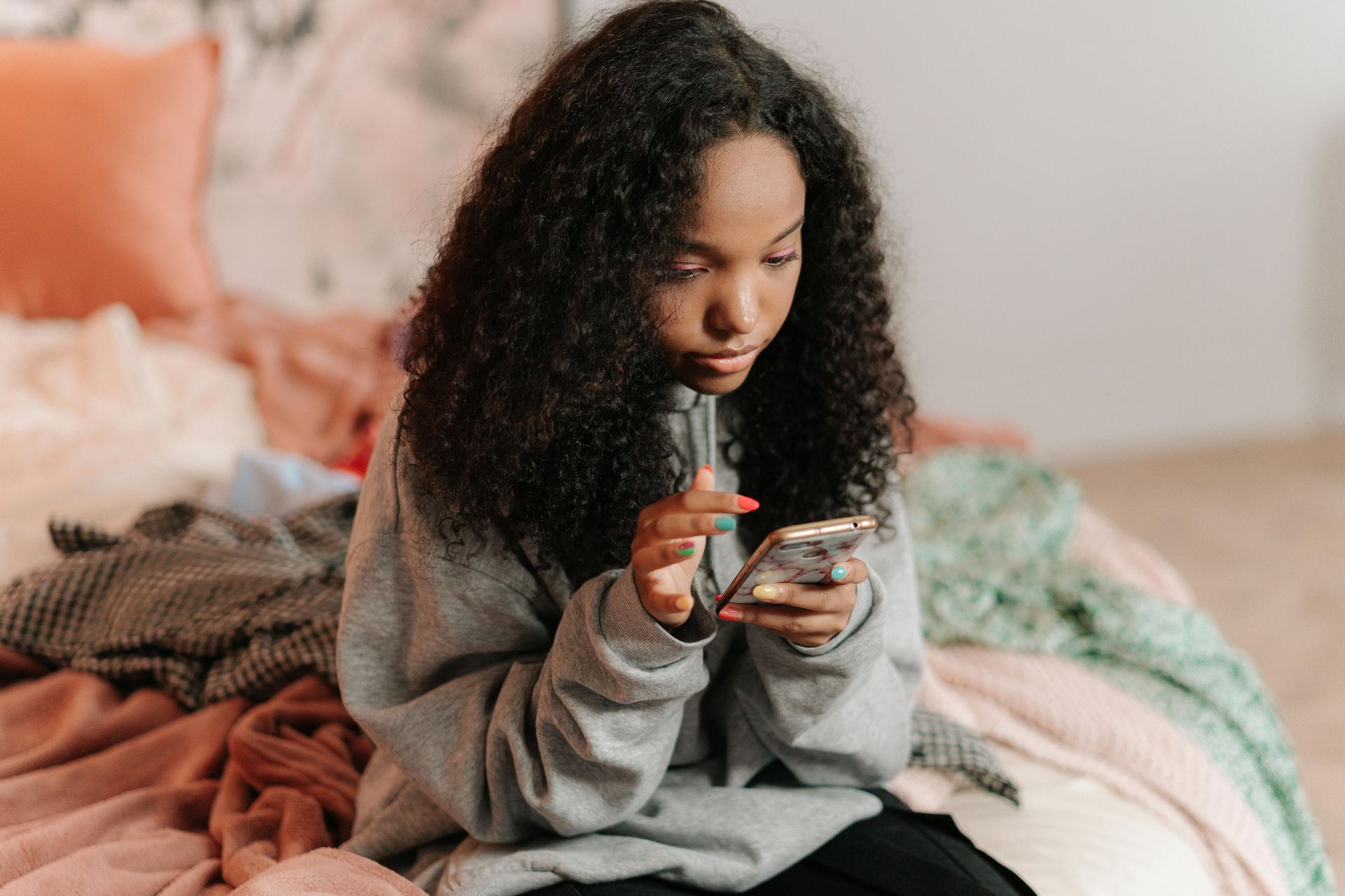 Person in gray sweatshirt using a phone while sitting on a bed with colorful blankets.