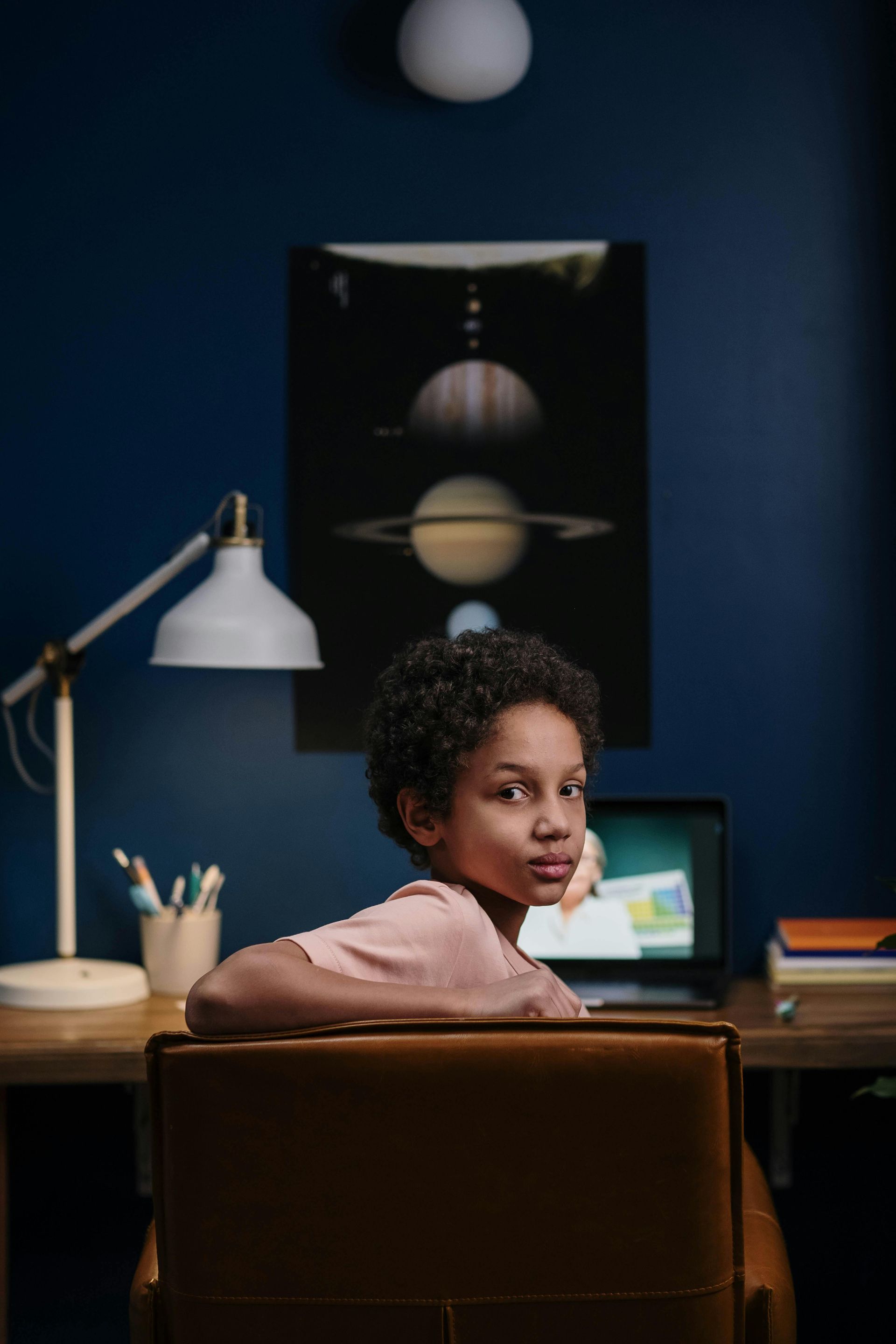 Girl sitting at desk with laptop, looking over shoulder. Dark blue wall, solar system poster, lamp.