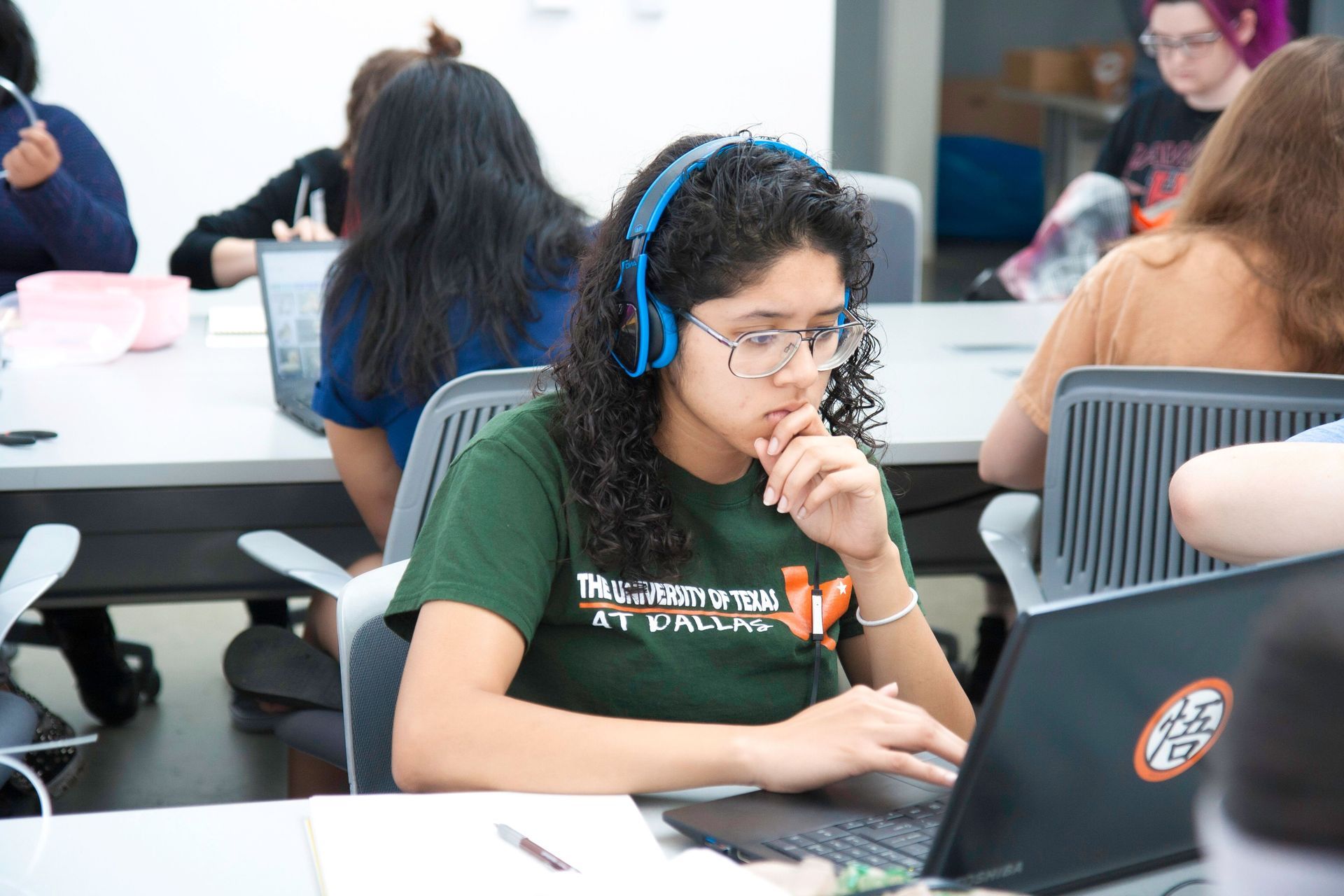 Student wearing blue headphones works on a laptop in a classroom.