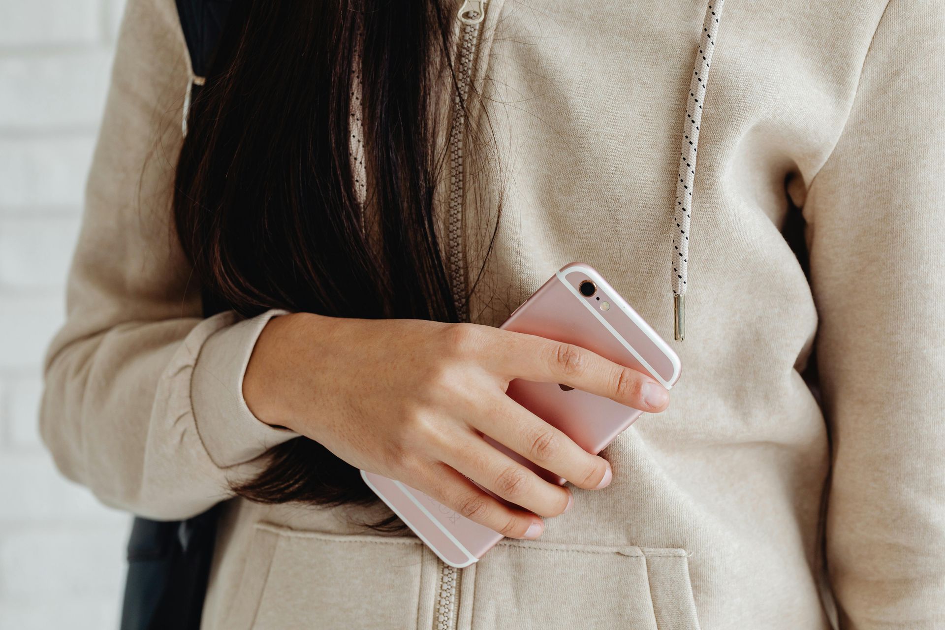 Person holding a rose gold smartphone, wearing a tan hoodie, against a white brick wall.