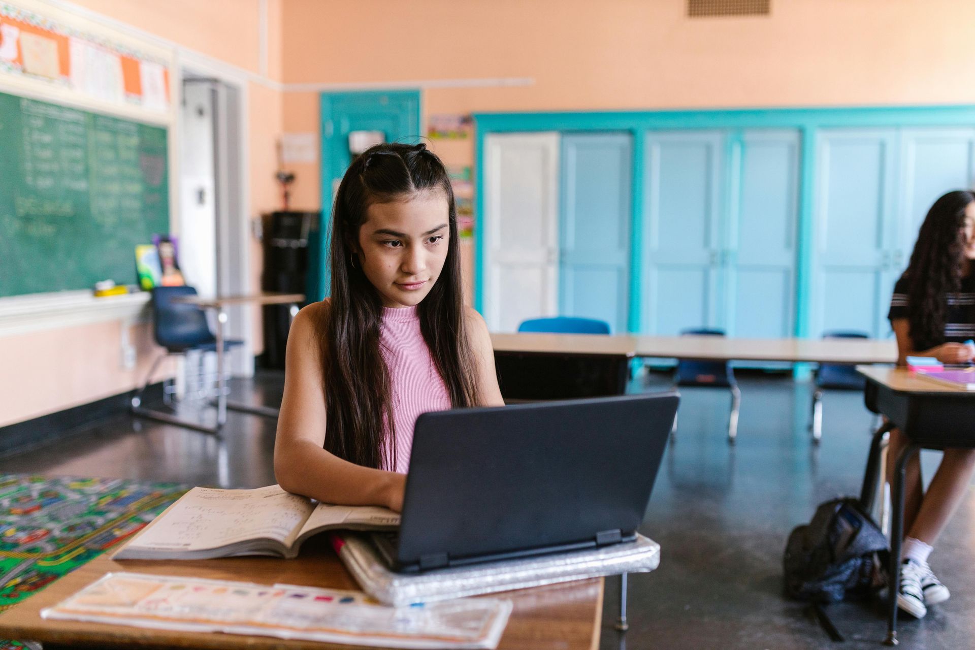 Girl with long hair using a laptop at a desk in a classroom, looking focused.