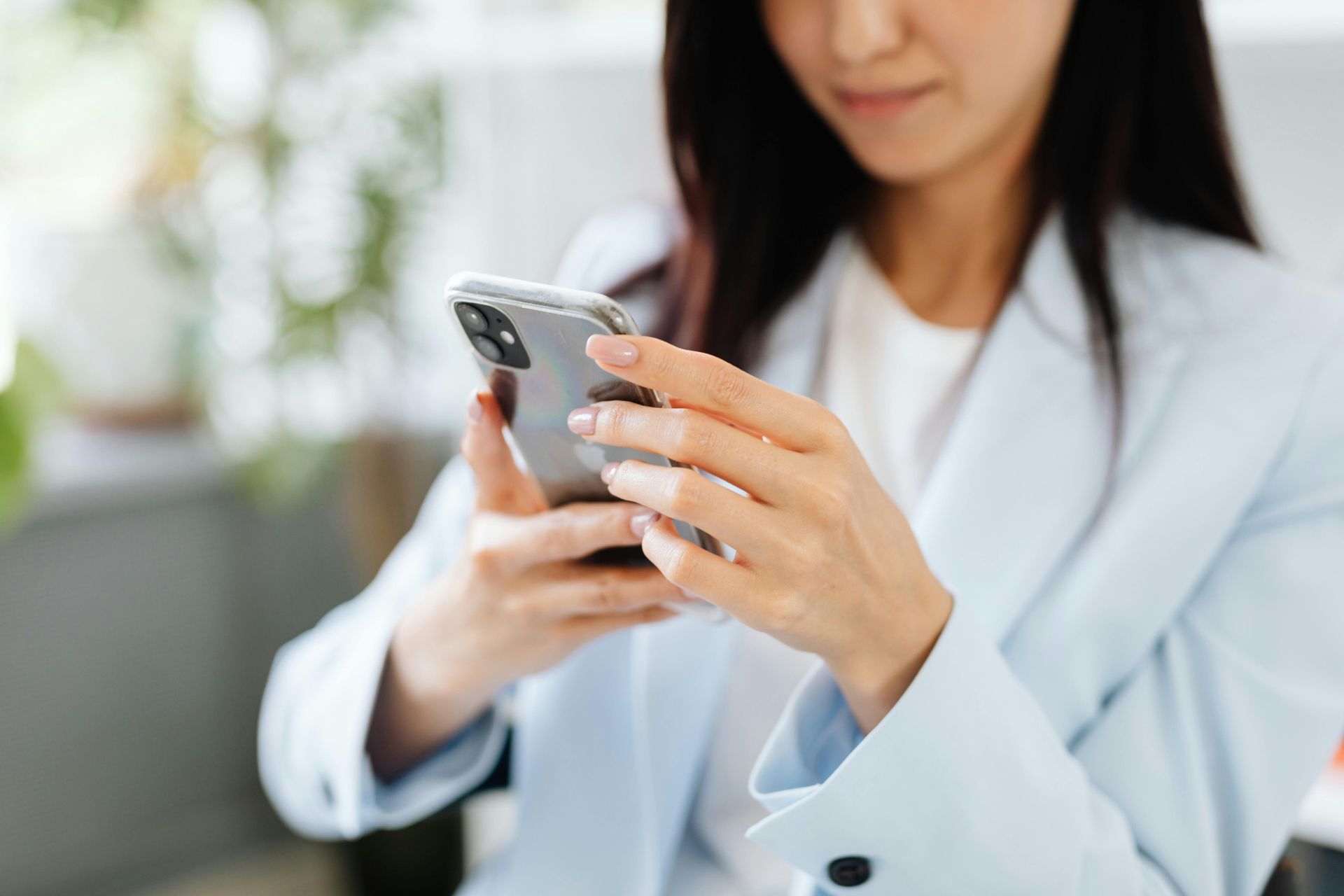 Woman in a light blue blazer using a smartphone indoors; blurred background.
