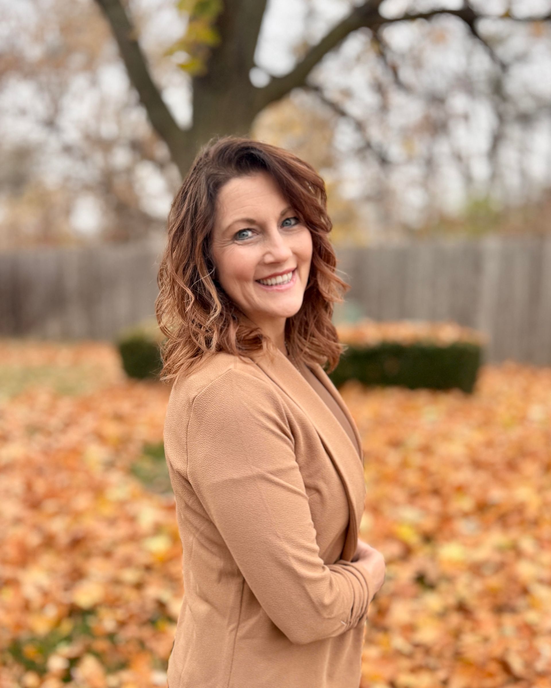 Woman in a tan jacket smiles outdoors in front of a tree and fallen leaves.