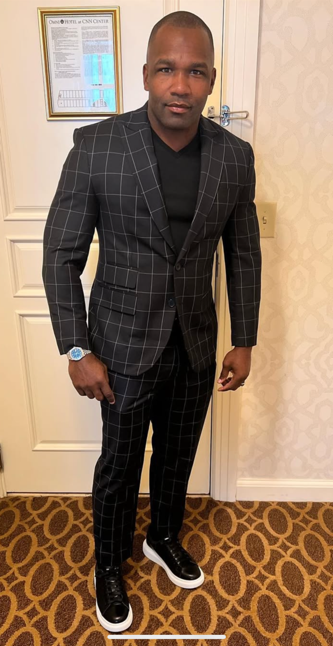 Man in black plaid suit, black shirt, and sneakers stands by a door indoors.