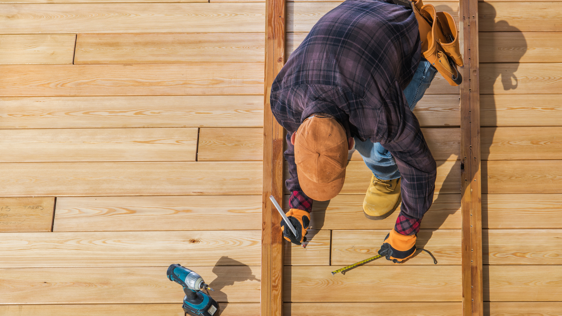 A man is working on a wooden deck with a drill and a tape measure.