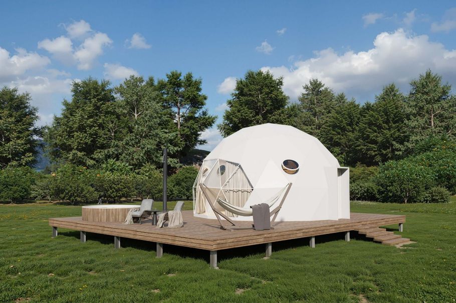 White dome-shaped glamping tent on a wooden deck with hammock, chairs, and hot tub; trees and sky in background.
