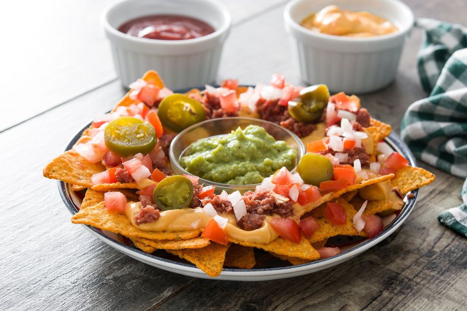 Nachos with toppings like salsa, guacamole, and jalapeƱos. Dips of red sauce and cheese sauce in the background.
