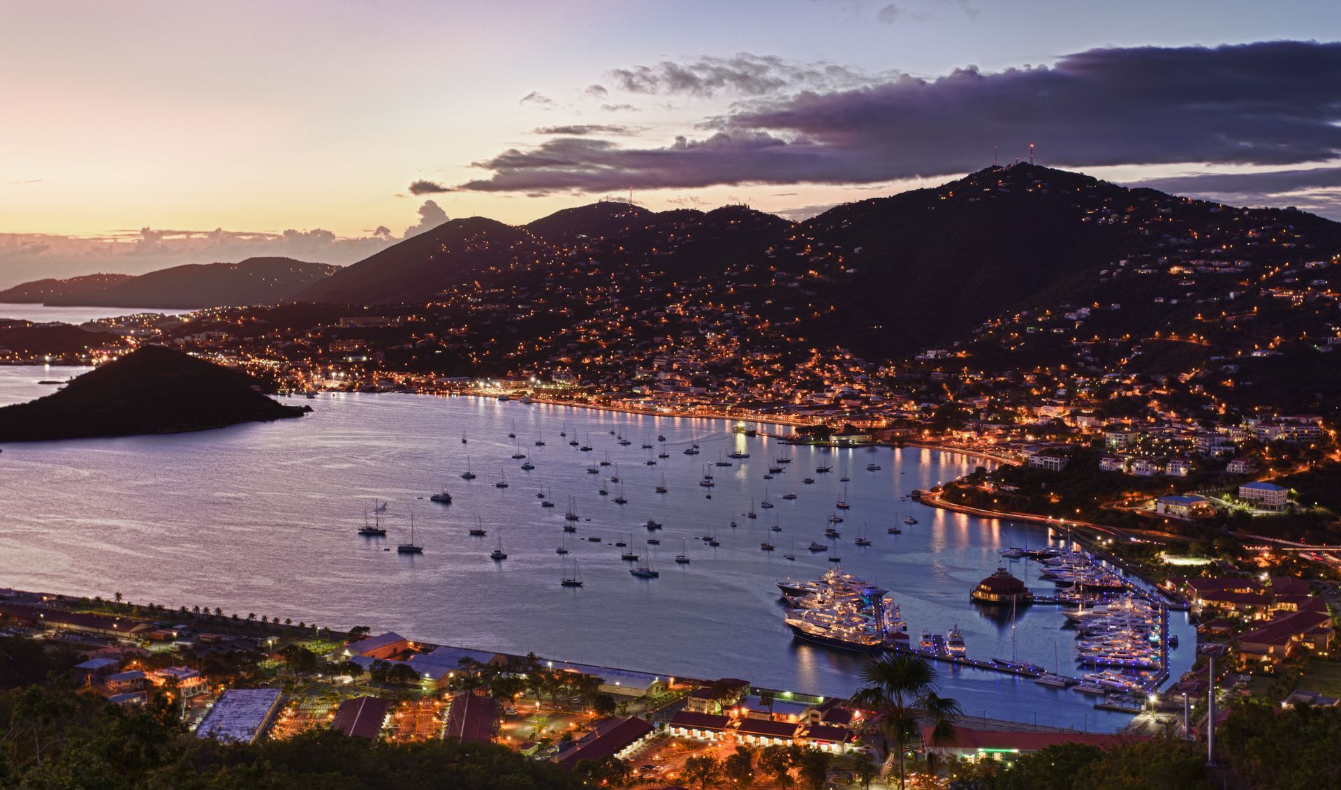 Night view of a harbor with numerous boats, city lights, and silhouetted hills under a dusky sky.