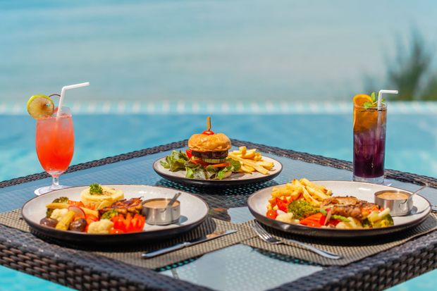Table set with food and drinks overlooking a pool. Dishes include a burger, fries, and entrees, plus colorful cocktails.