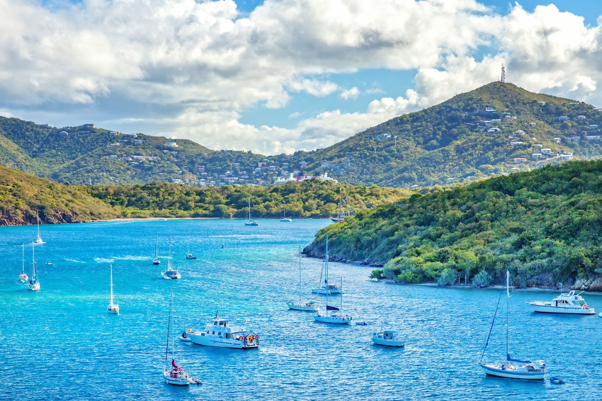 Sailboats in a turquoise bay, surrounded by lush, green hills under a partly cloudy sky.