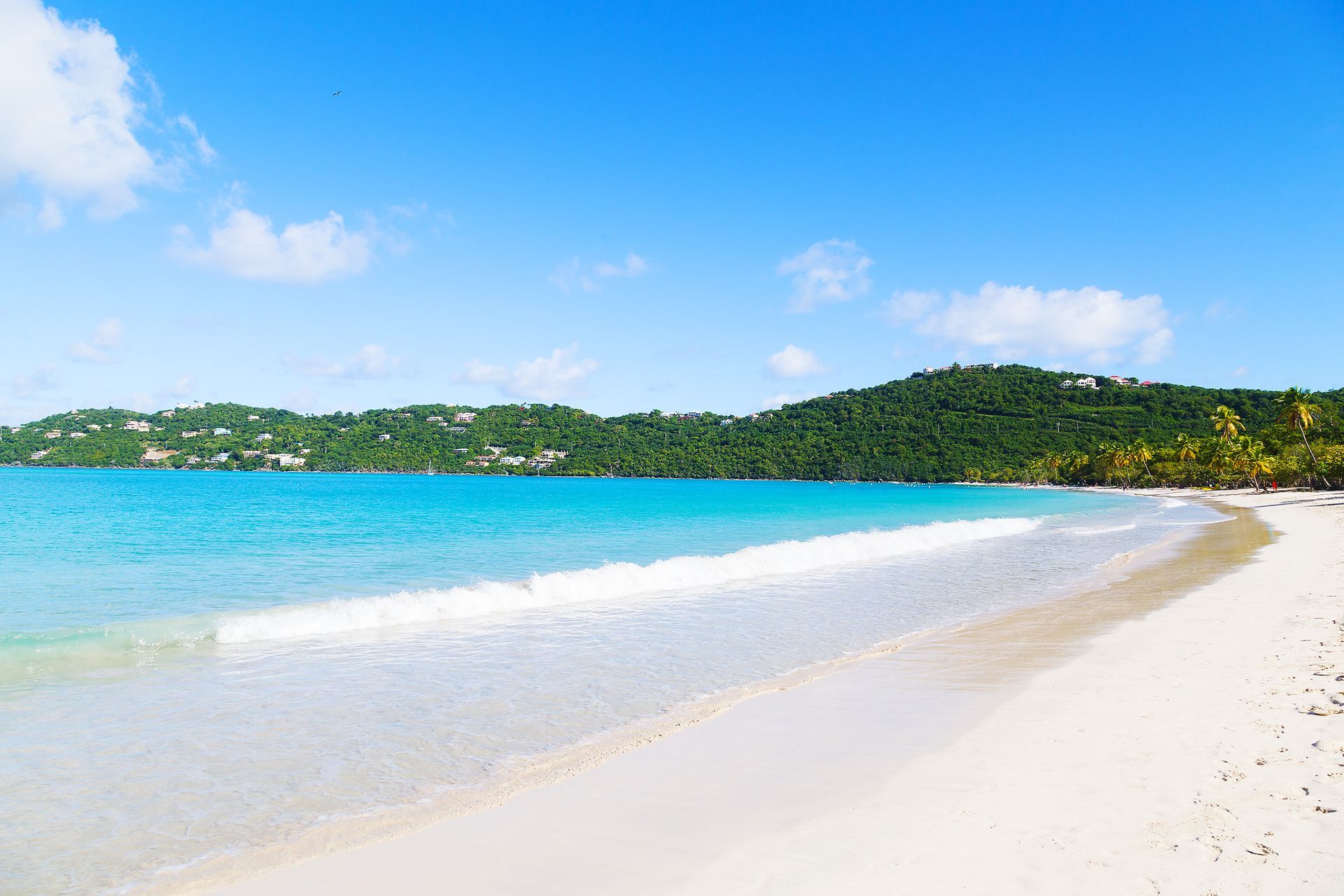 White sandy beach with turquoise water under a blue sky, hills in the background.