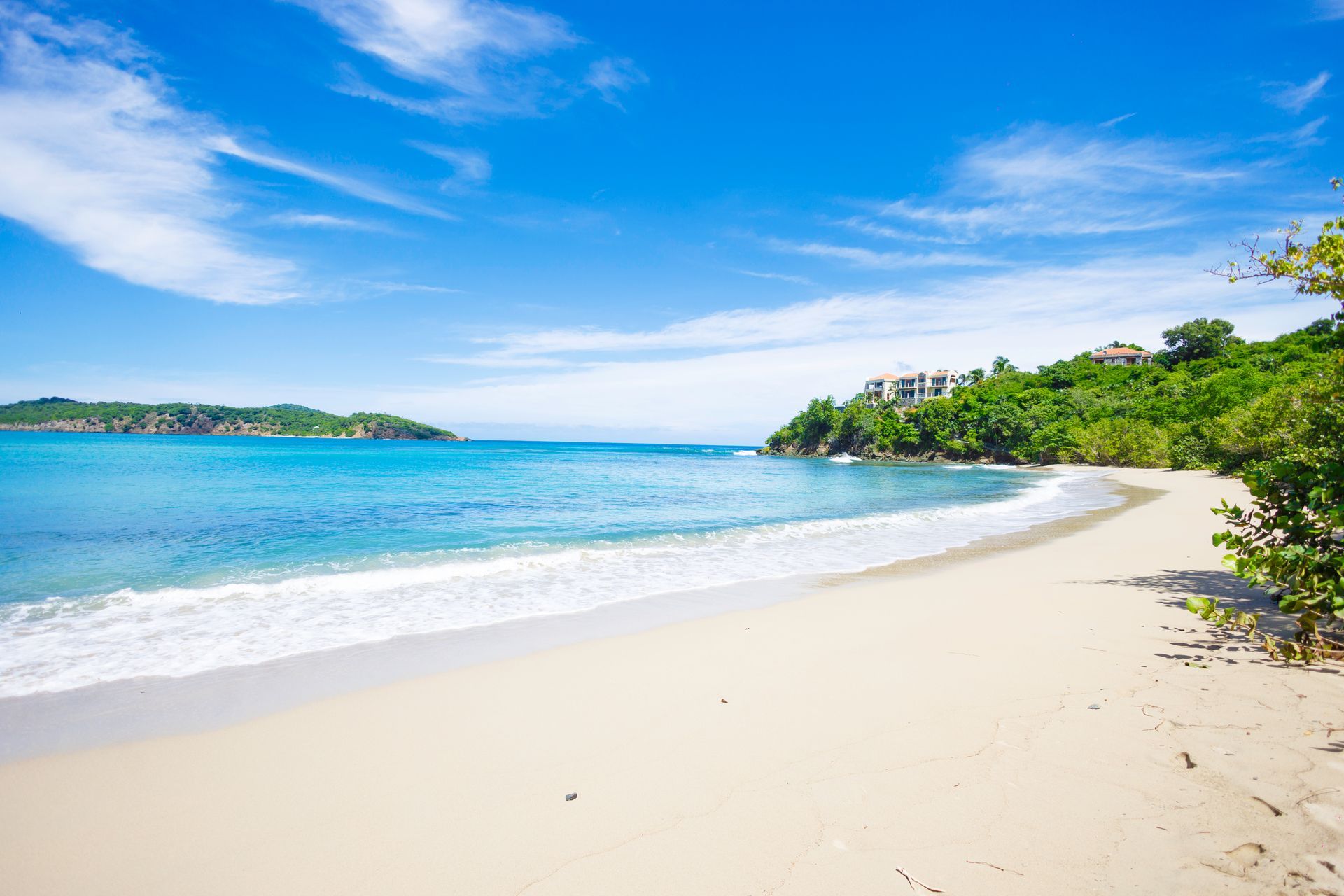 Sandy beach with clear turquoise water and blue sky, with greenery and a small island in the background.