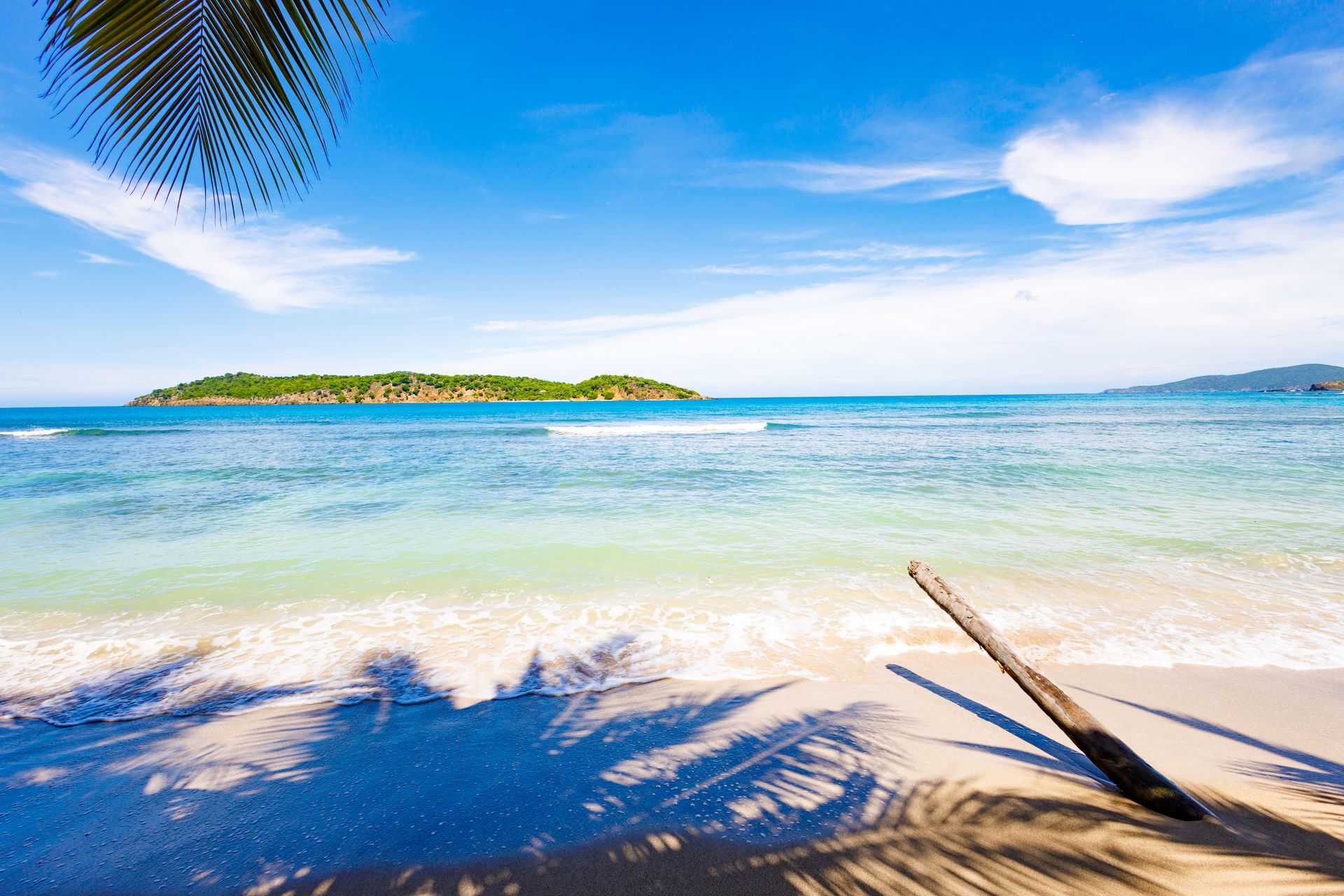 Beach scene with clear blue water, white sand, and small island in the distance under a bright sky.